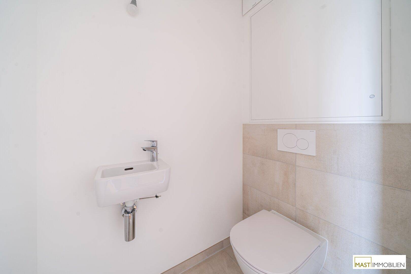 Separates WC mit wandhängender Toilette und kleinem Handwaschbecken, halbhoch gefliest.