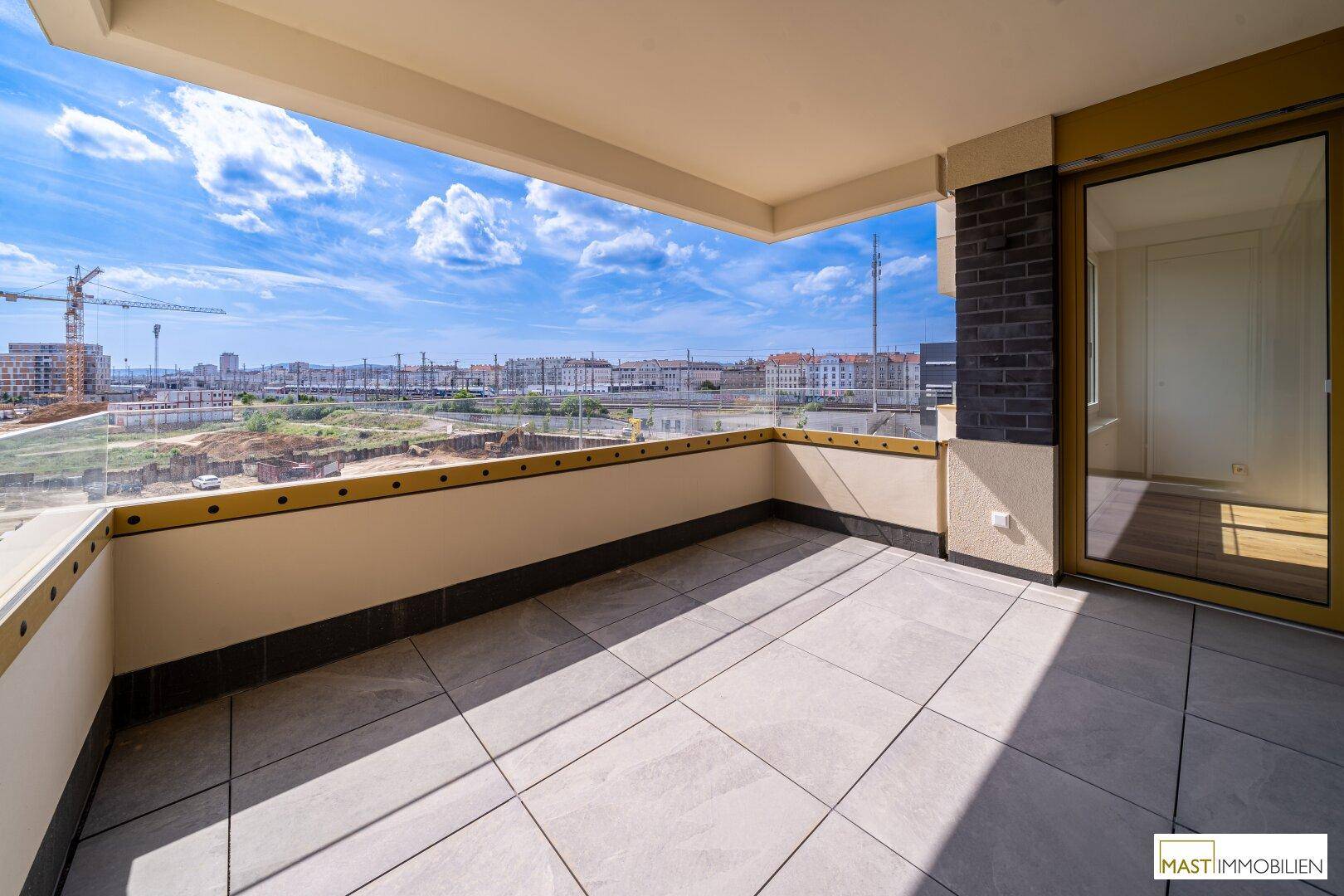 Geräumiger Balkon mit Fliesenboden und Glasgeländer, bietet einen weiten Blick über die Stadt und eine Baustelle.