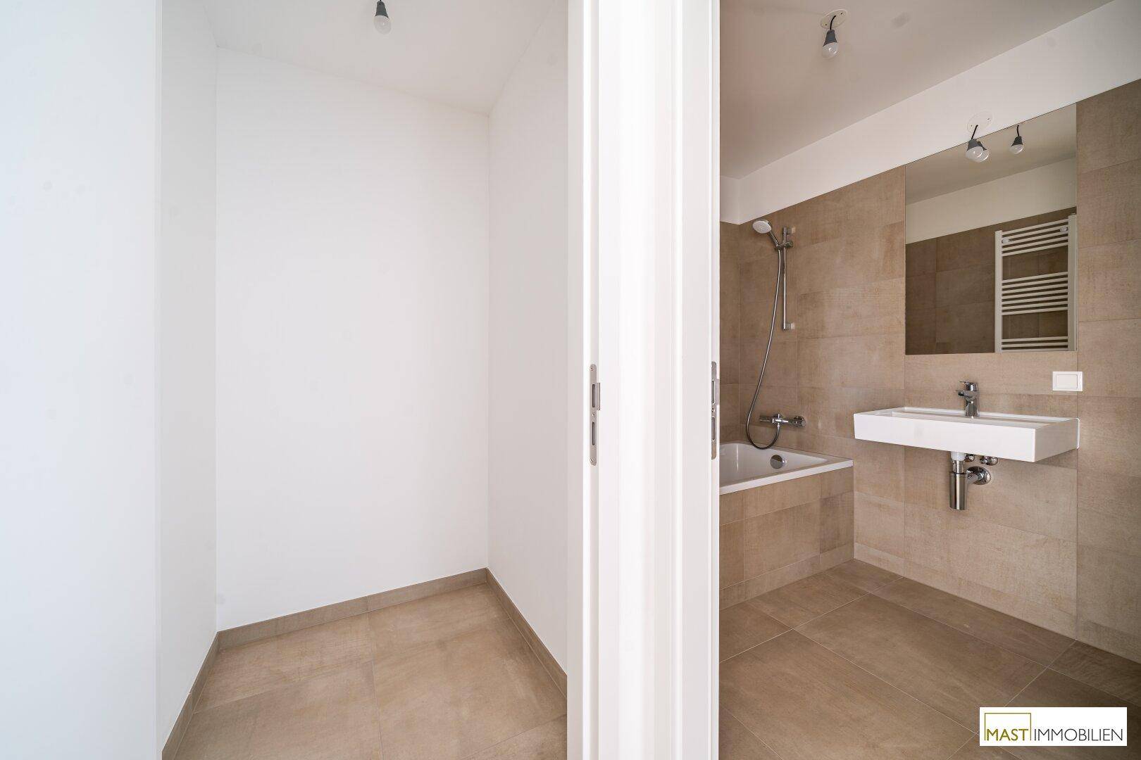 Modernes Badezimmer mit Badewanne, Waschbecken und beigefarbenen Fliesen, angrenzend an einen leeren Raum.