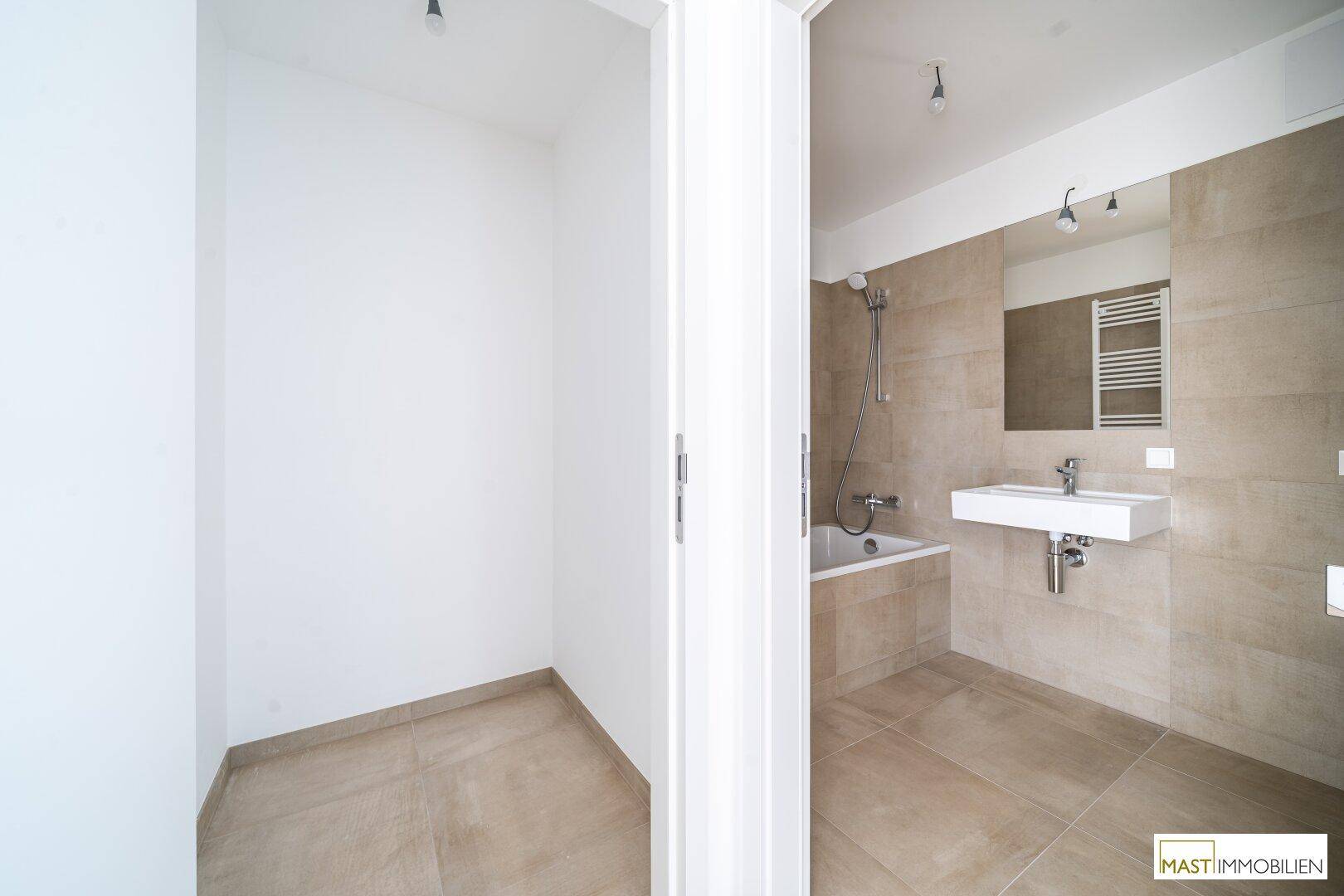 Modernes Badezimmer mit Badewanne, Waschbecken und beigefarbenen Fliesen, angrenzend an einen Abstellraum.