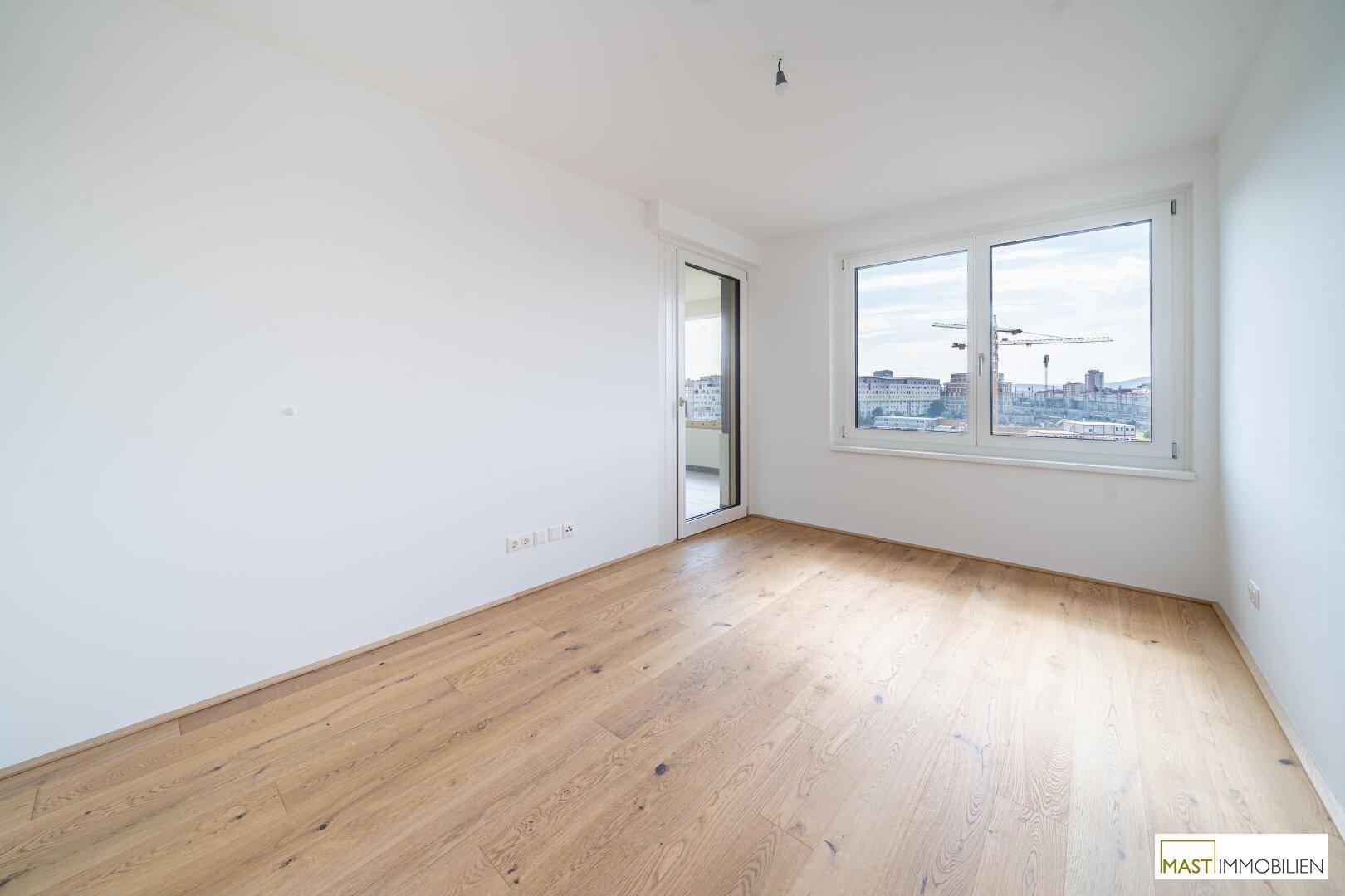 Heller, leerer Raum mit Holzboden und großen Fenstern, die einen Blick auf die Stadt ermöglichen.