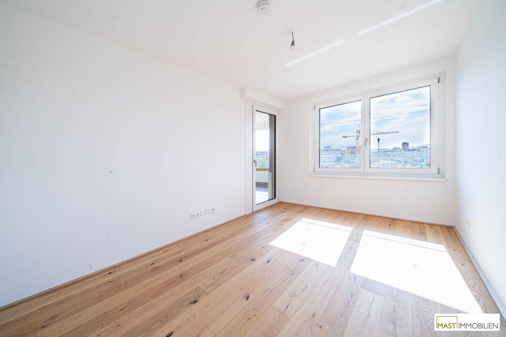 Heller, leerer Raum mit Holzboden und großen Fenstern, die viel Tageslicht hereinlassen.