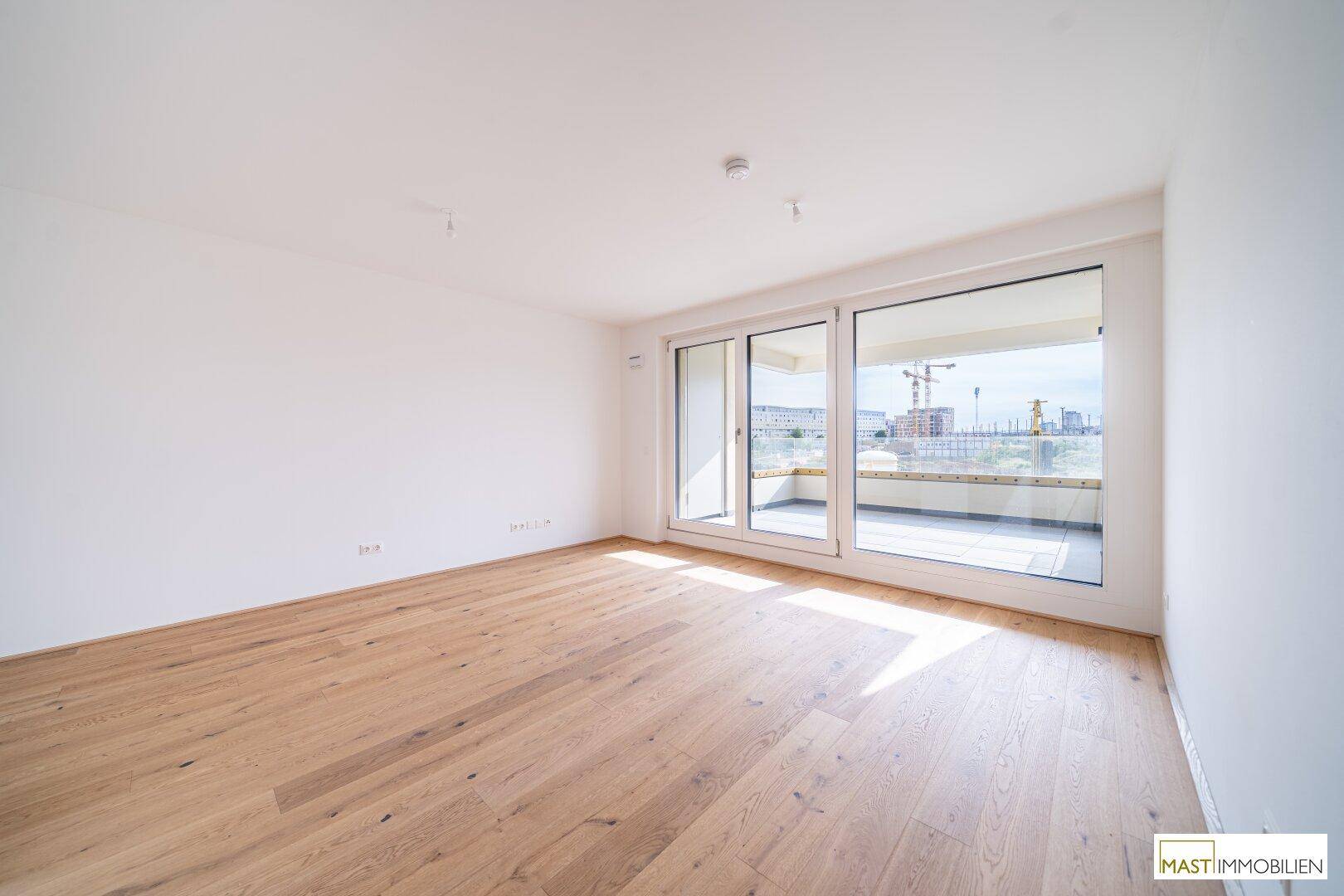 Helles, leeres Zimmer mit Holzboden und großen Fenstern, die viel Tageslicht hereinlassen.