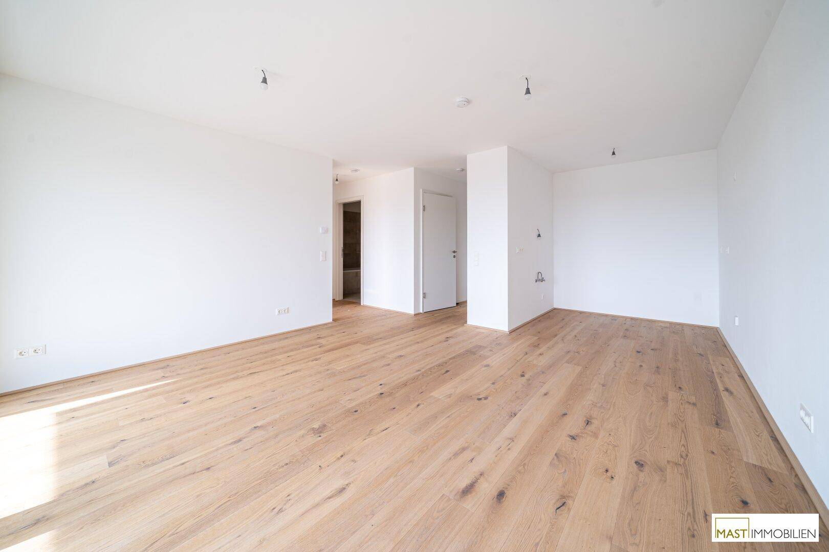 Geräumiger Wohnbereich mit hellem Holzboden und weiß gestrichenen Wänden, bietet viel Gestaltungsfreiheit.