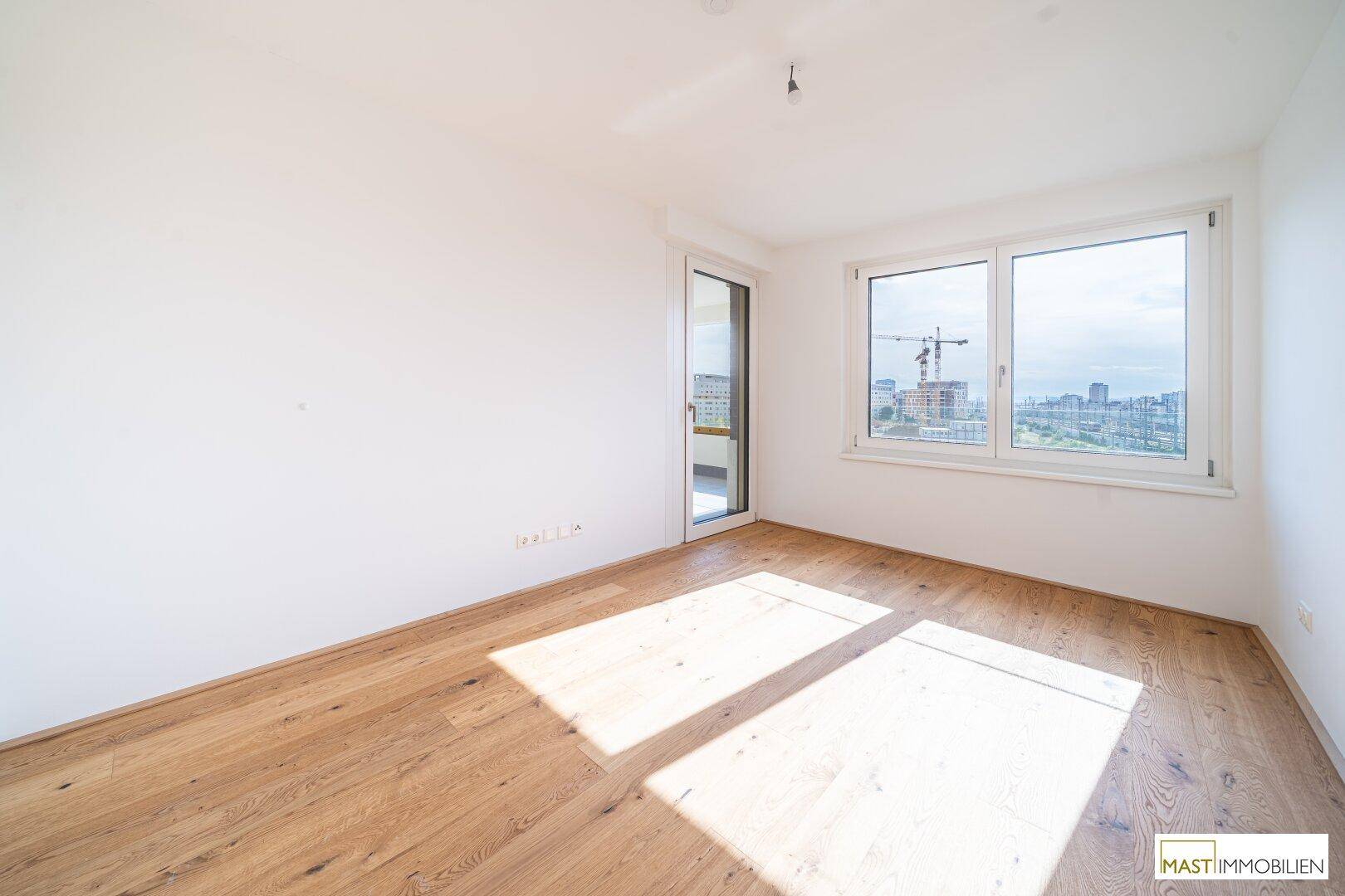 Heller, leerer Raum mit Holzboden, großen Fenstern und Zugang zu einem Außenbereich.