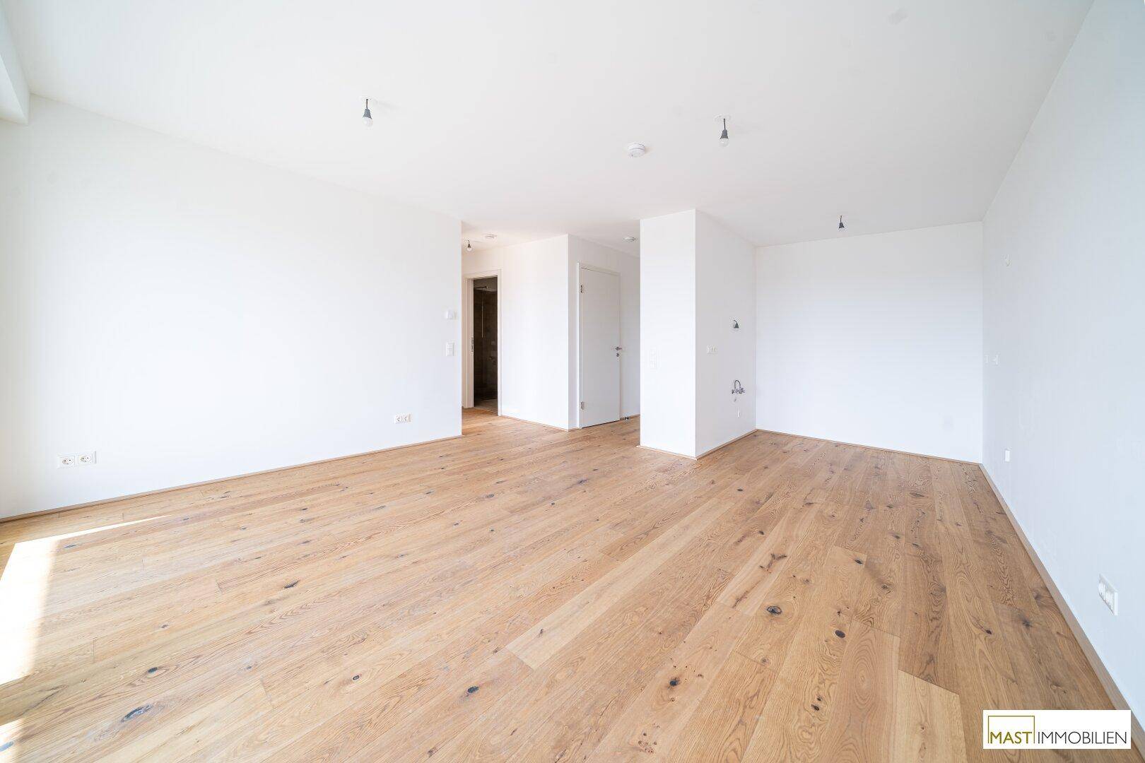 Heller, leerer Raum mit Holzboden und weiß gestrichenen Wänden, offen zum Flur.