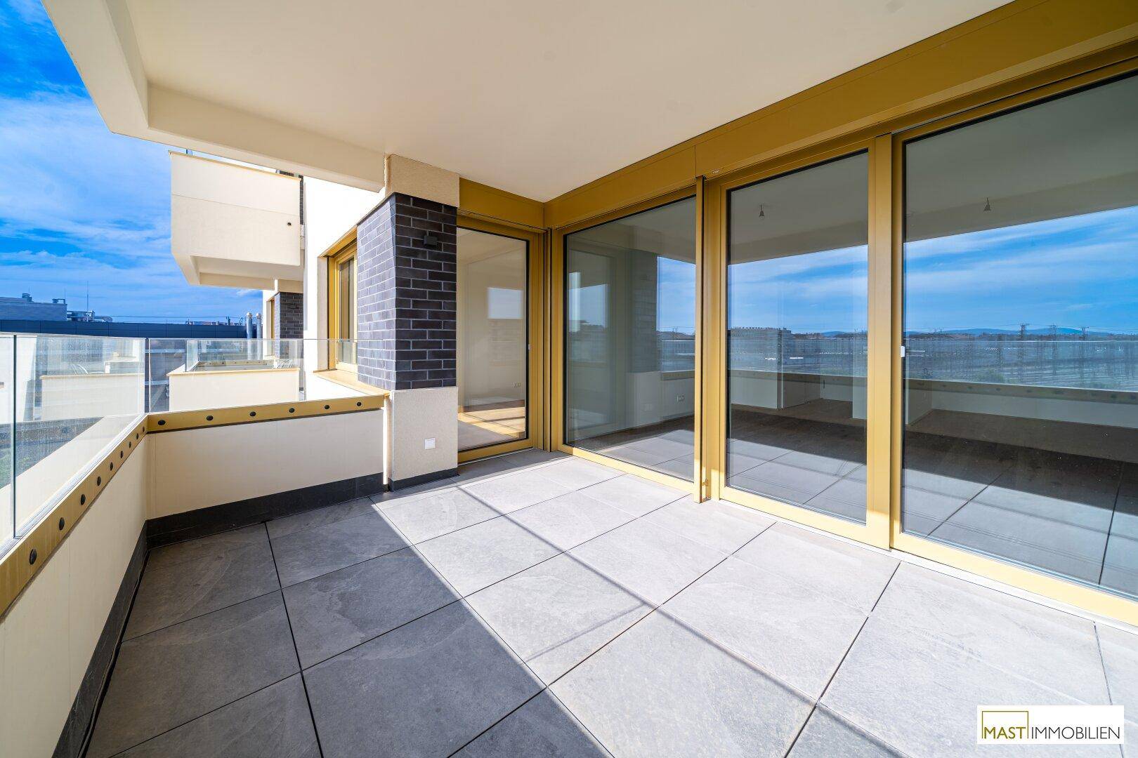 Weitläufige, geflieste Terrasse mit Glasgeländer und Blick auf die Umgebung und den Himmel.