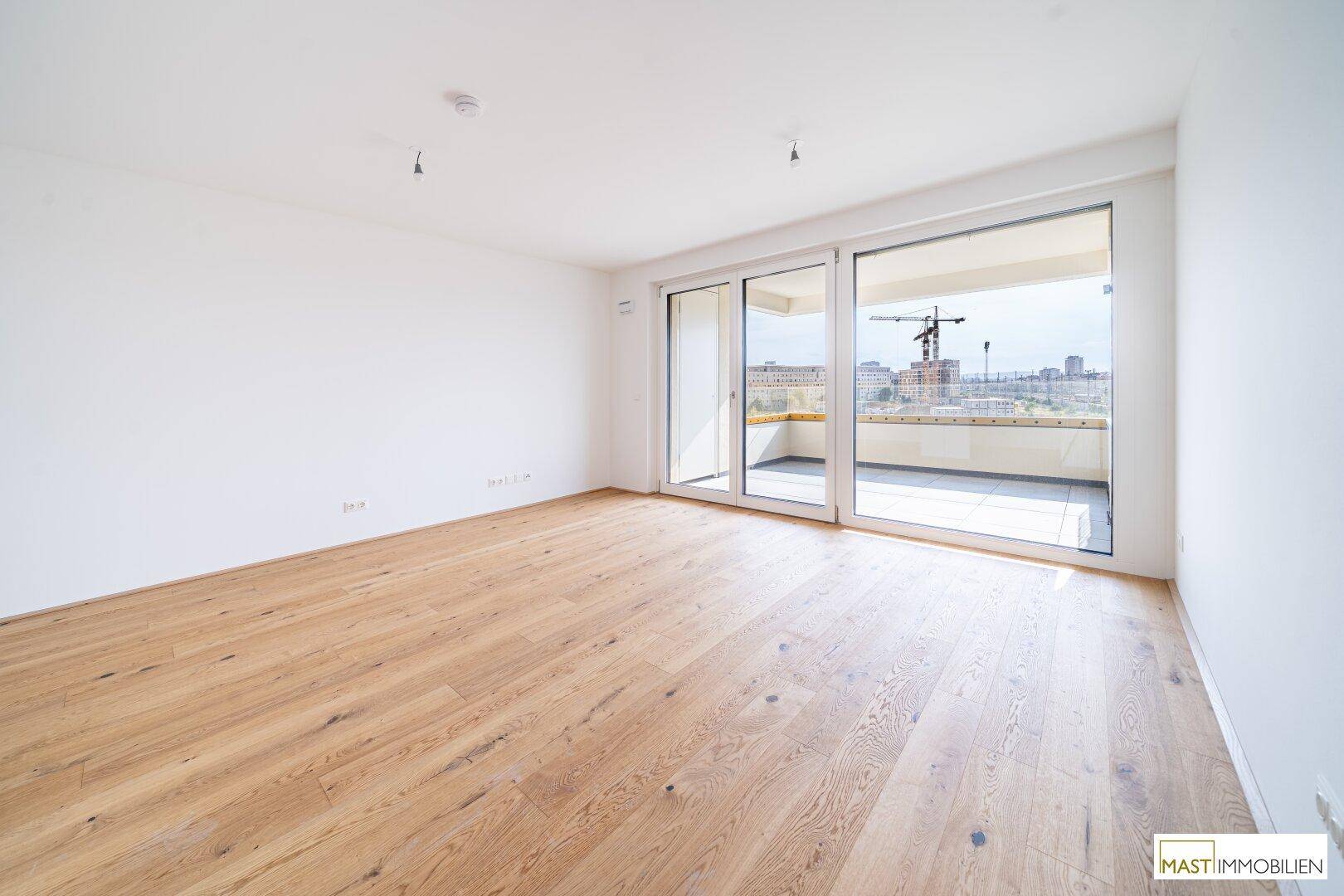 Geräumiges Wohnzimmer mit Parkettboden und Zugang zu einem Balkon mit Blick auf die Stadt und Baustellen.