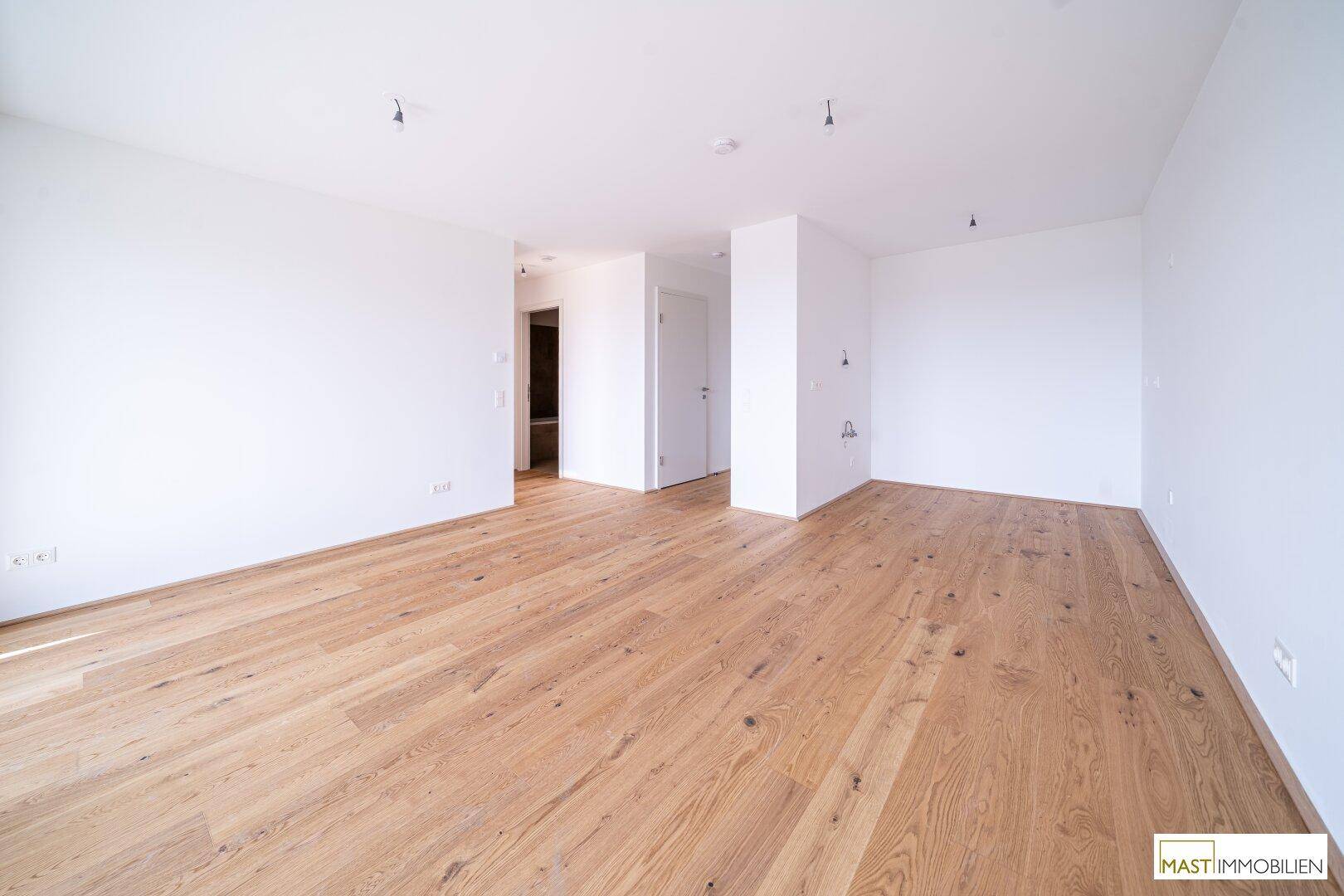 Heller, leerer Raum mit Holzboden und weißen Wänden, der einen offenen Übergang zu einem Flur zeigt.