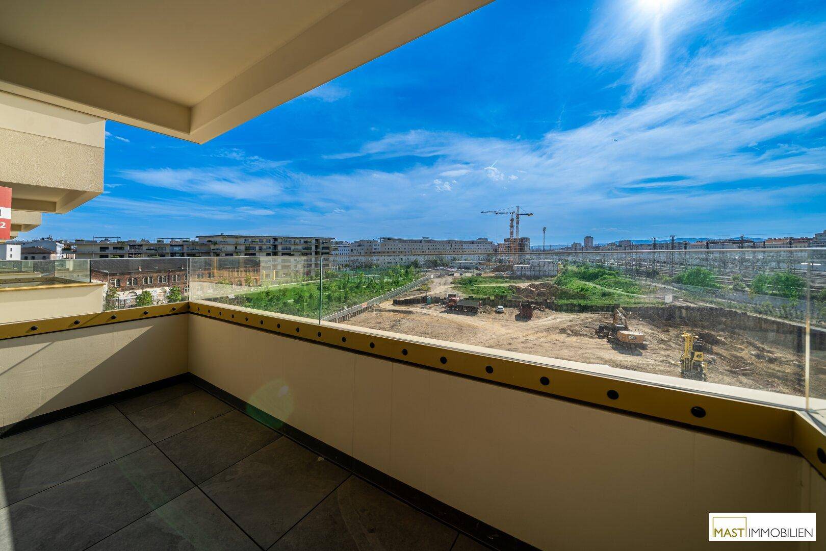 Sonniger Balkon mit Fliesenboden und Glasgeländer, bietet einen weiten Blick auf Baustellen und die Stadt.