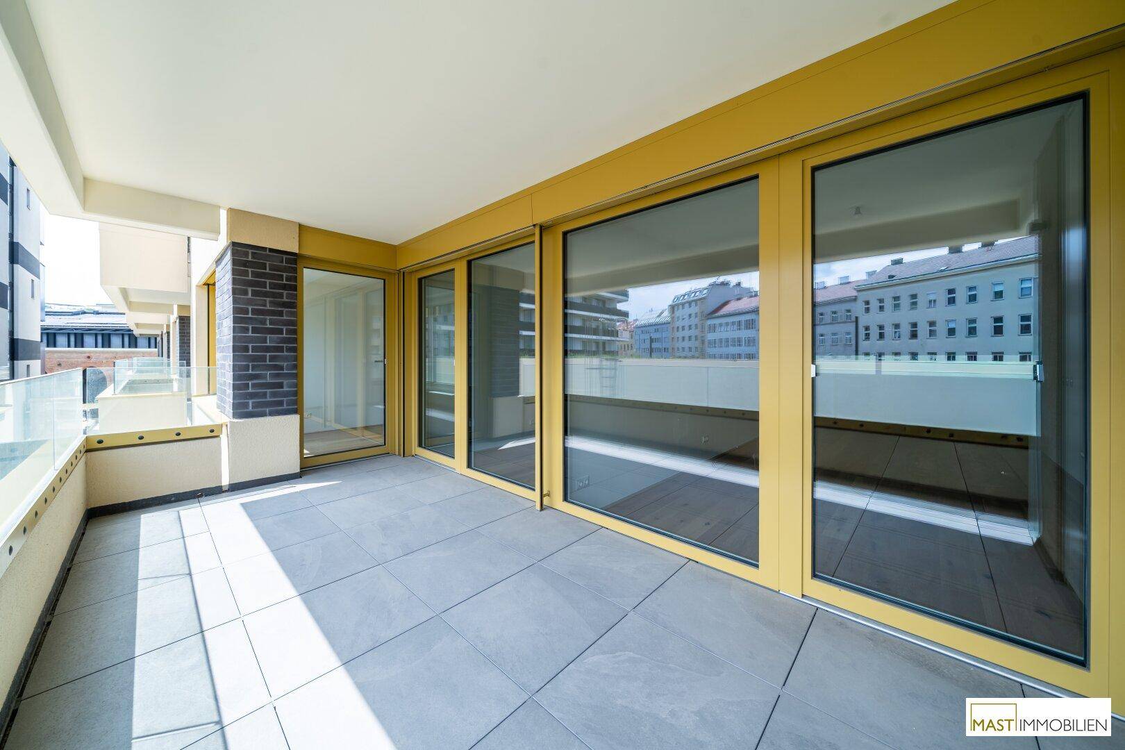 Geräumiger Balkon mit Fliesenboden, Glasgeländer und Blick auf die umliegenden Gebäude.