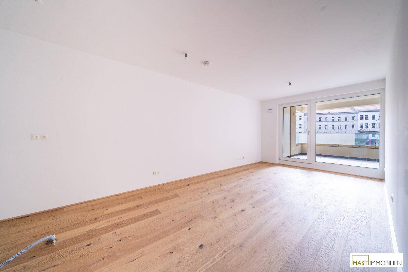 Geräumiger, heller Raum mit Holzboden und Zugang zu einem Balkon, der viel Tageslicht hereinlässt.