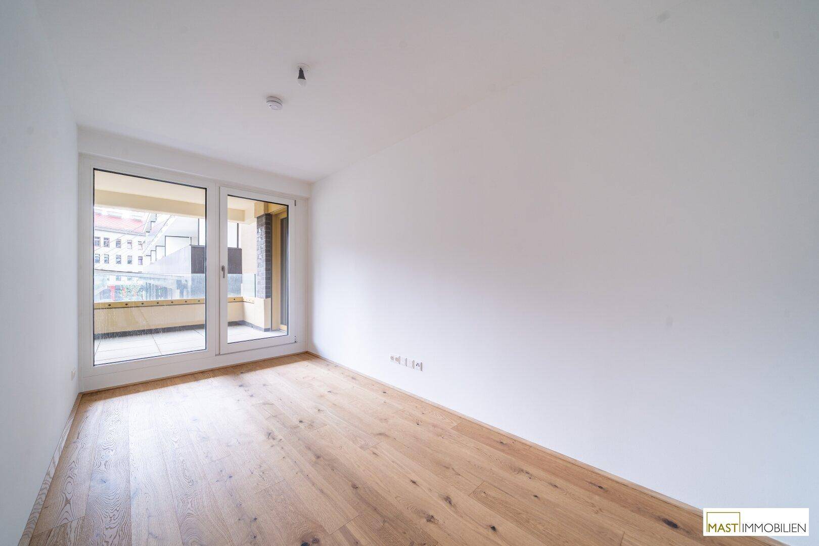 Leerer Raum mit Holzboden und großen Fenstern, die den Zugang zu einem Außenbereich ermöglichen.
