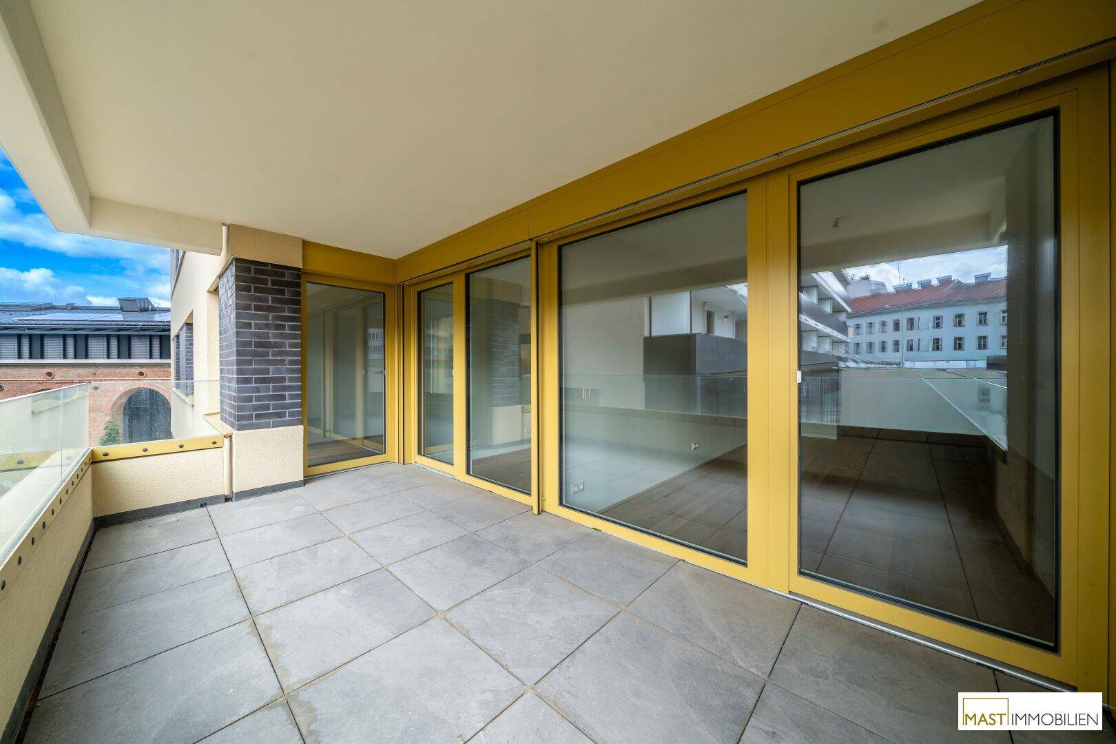 Geräumige Terrasse mit Fliesenboden und gelben Fensterrahmen, die einen hellen Außenbereich bietet.