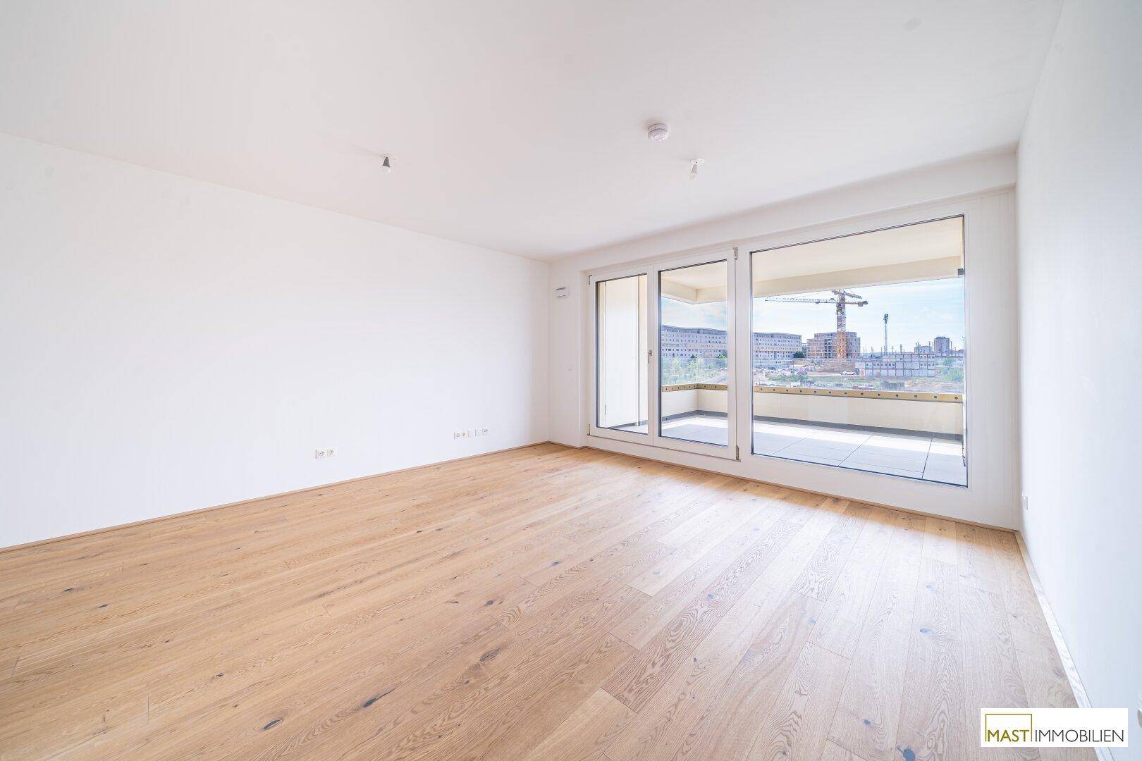 Heller, leerer Raum mit Holzparkettboden und Zugang zu einem sonnigen Balkon.