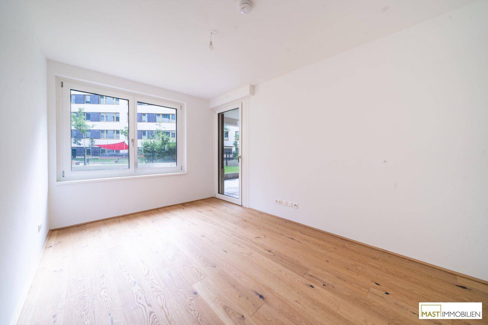 Helles Schlafzimmer mit Parkettboden und Zugang zu einem Außenbereich durch Fenster und Tür.