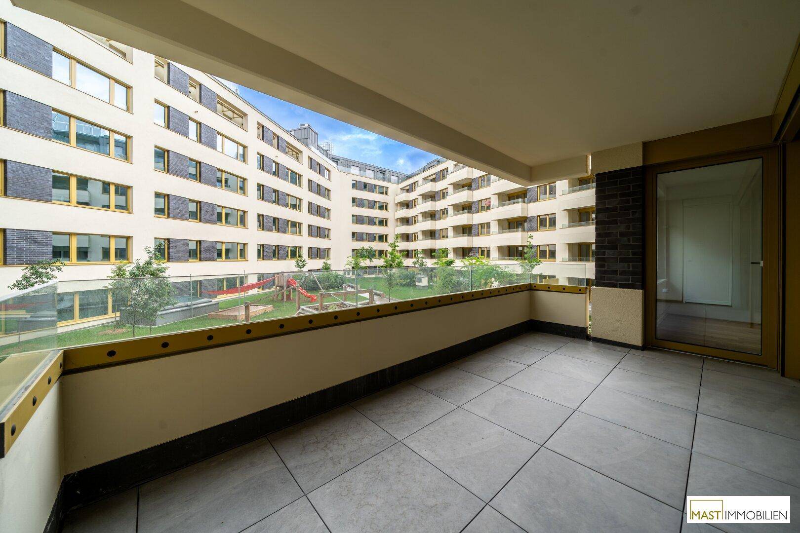 Geräumiger Balkon mit Fliesenboden und Glasbrüstung, mit Blick auf den begrünten Innenhof.