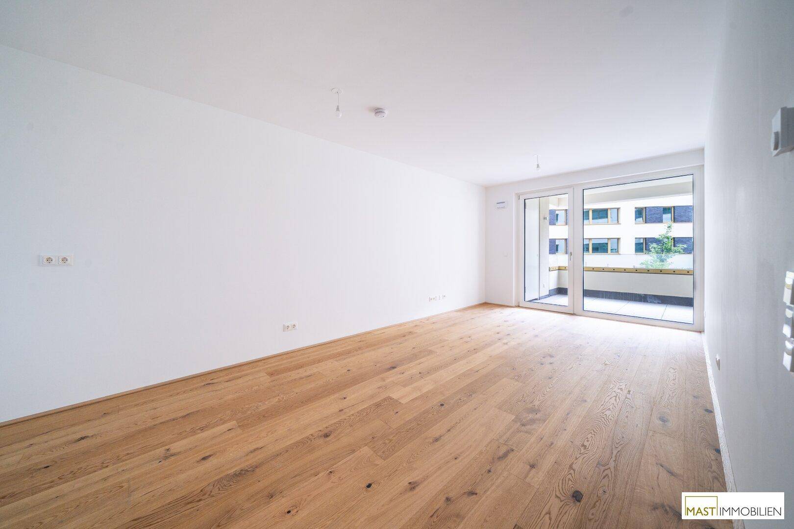 Geräumiges Wohnzimmer mit hellem Parkettboden und großen Fenstern, die viel Tageslicht hereinlassen.