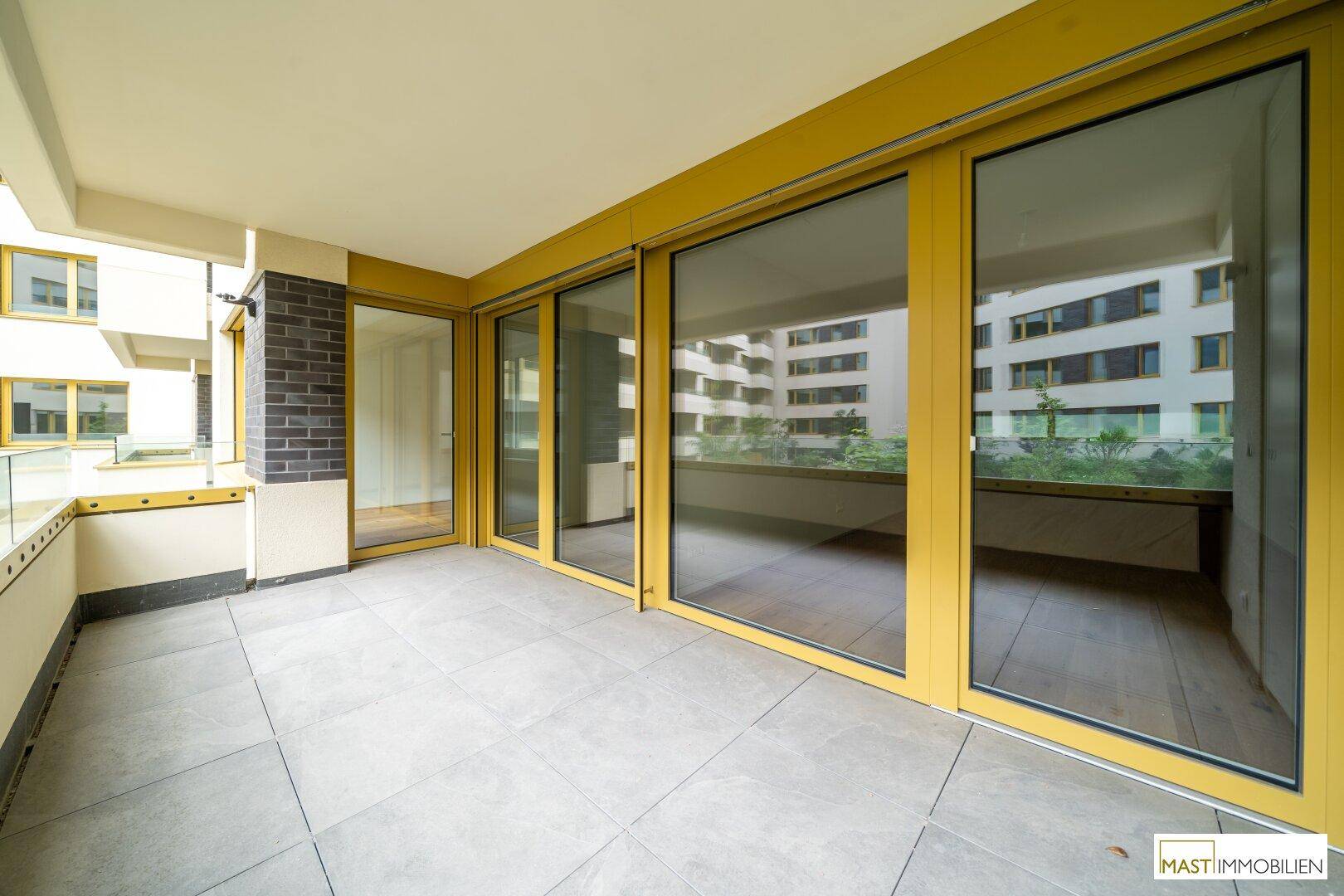 Geräumiger Balkon mit Fliesenboden und gelben Fensterrahmen, bietet einen Blick auf die umliegenden Gebäude.