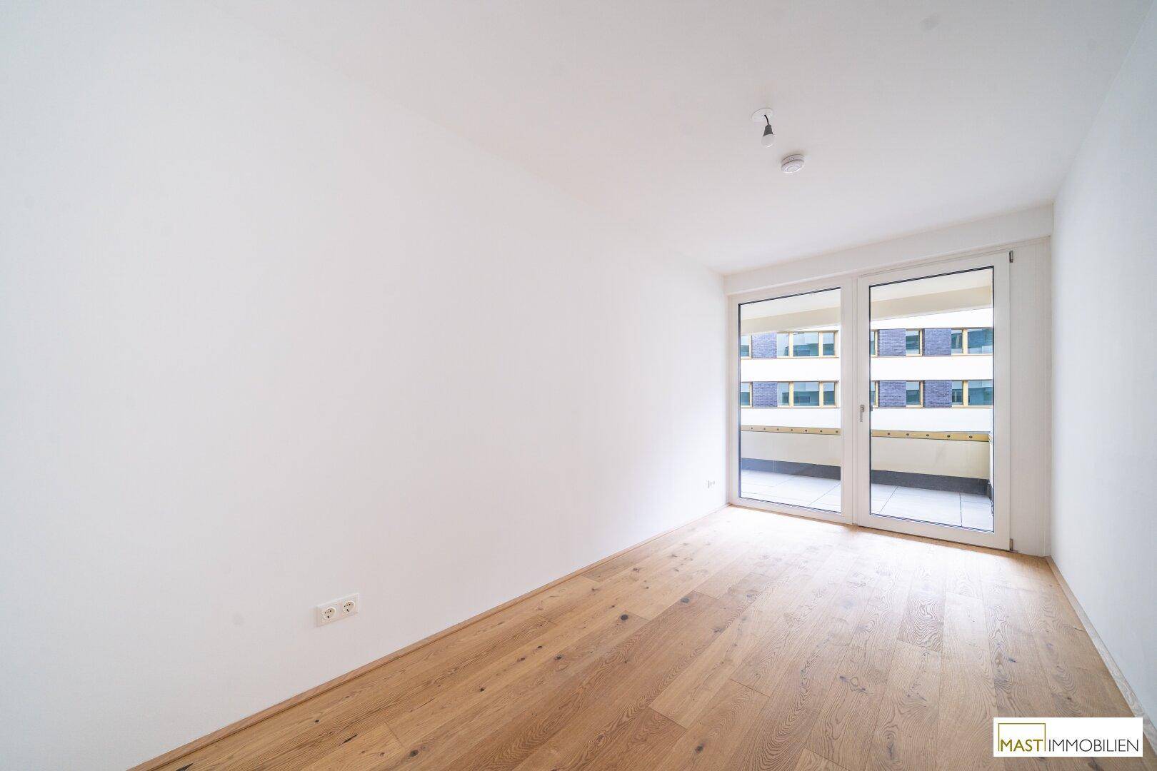 Helles Schlafzimmer mit Holzparkettboden und großen Fenstern, die Zugang zu einem Außenbereich bieten.