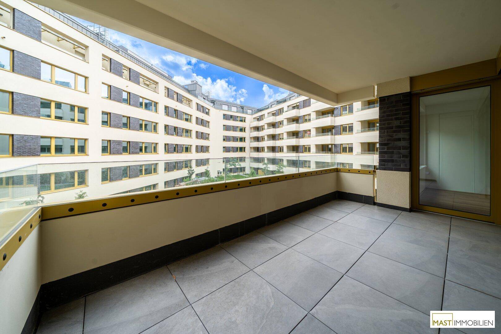 Großer Balkon mit Fliesenboden und Glasbrüstung, bietet einen weiten Blick auf den begrünten Innenhof.