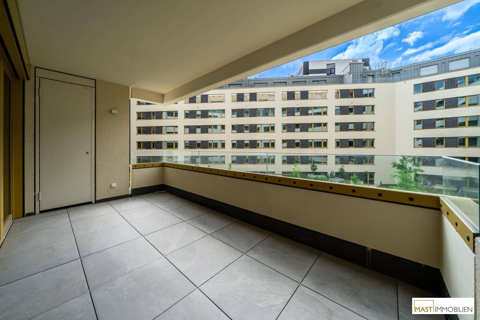 Geräumiger Balkon mit Fliesenboden und Glasgeländer, bietet einen Blick auf die umliegenden Gebäude.