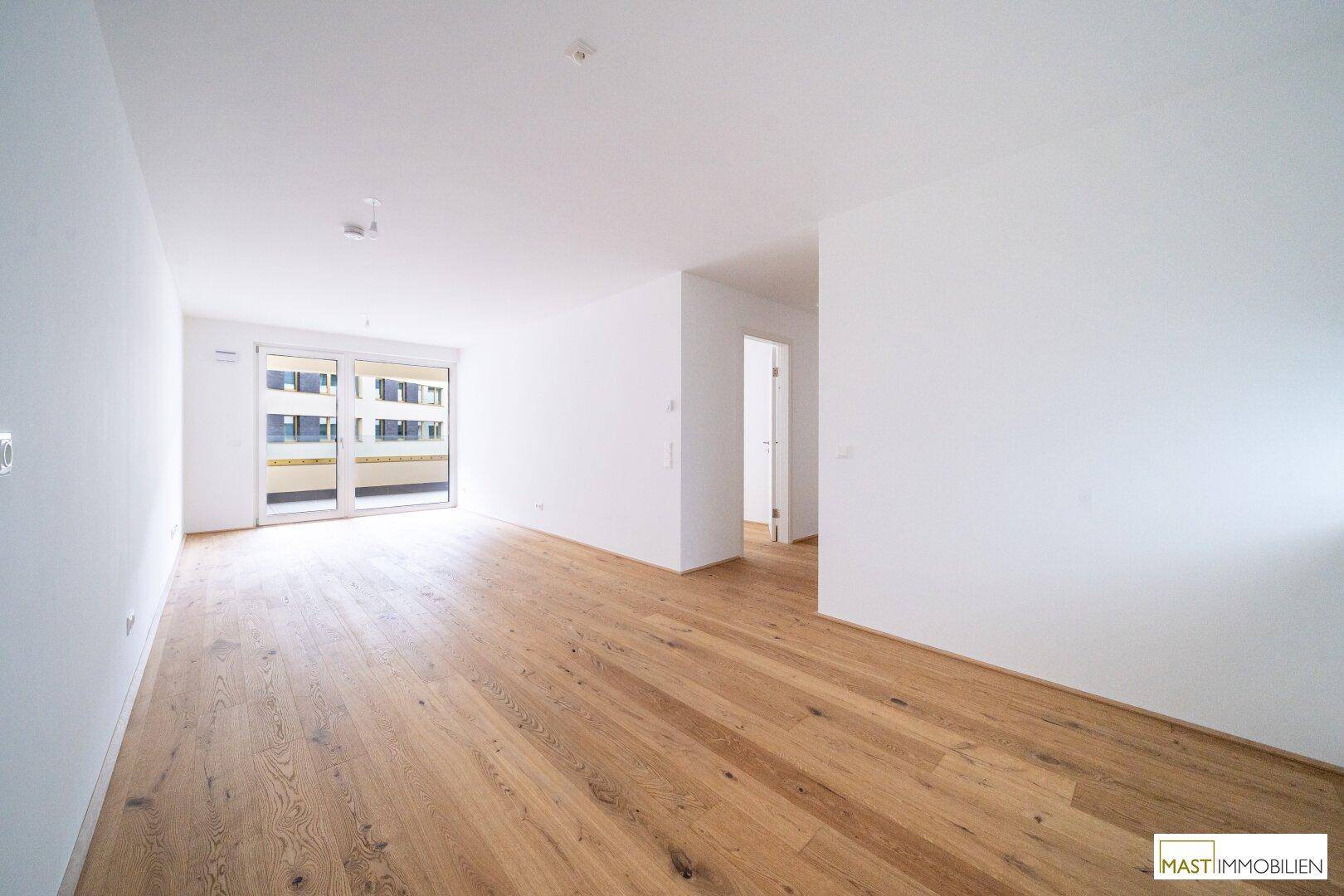 Offener Wohnbereich mit hellem Holzboden und Zugang zu einem Balkon, bietet viel Tageslicht.