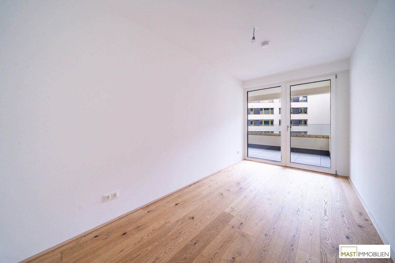 Helles, leeres Zimmer mit Eichenparkettboden und Zugang zu einem Balkon durch große Glastüren.