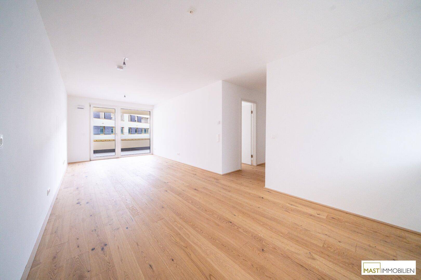 Geräumiges, leeres Zimmer mit hellem Eichenparkett und Zugang zu einem Balkon, ideal für offenes Wohnen.