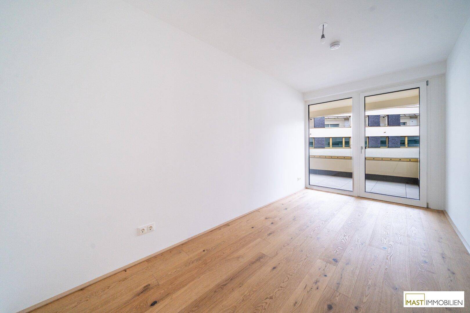 Helles Zimmer mit Eichenparkettboden und großen Fenstern, die auf einen Balkon führen und viel Licht spenden.