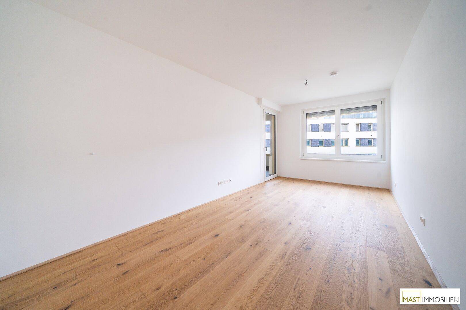 Leeres Zimmer mit Holzboden und großem Fenster, das viel Tageslicht hereinlässt.