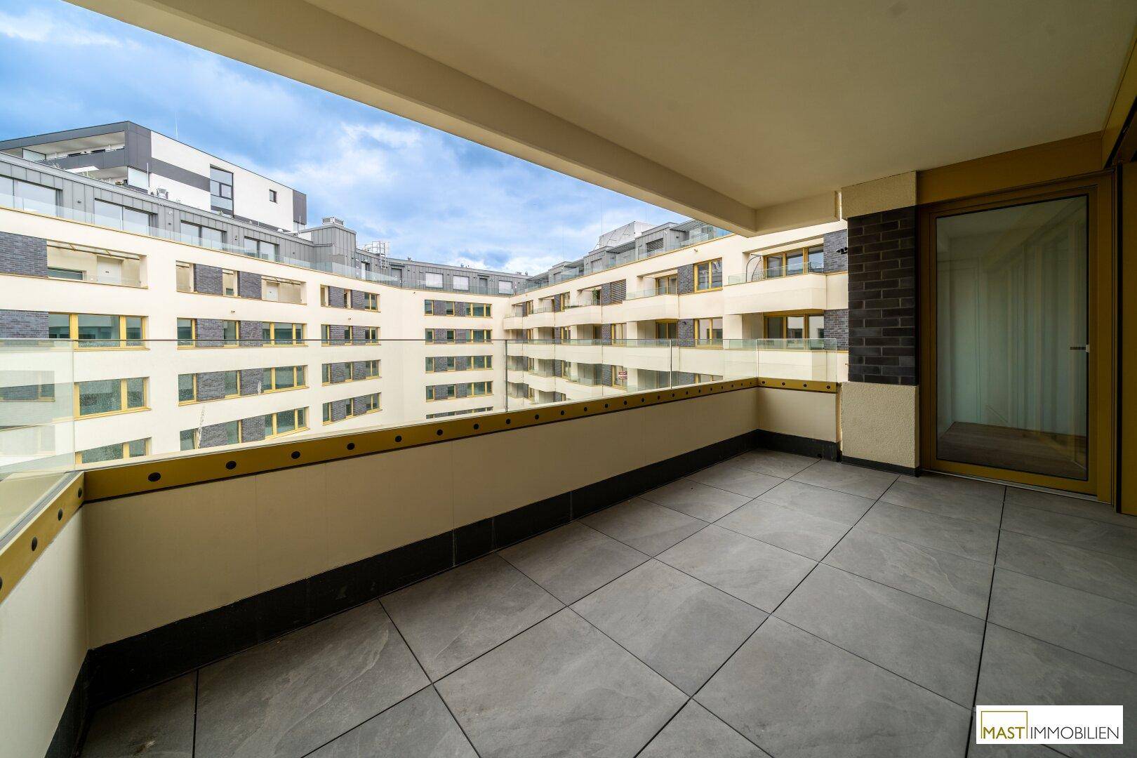 Geräumiger Balkon mit Fliesenboden und Glasgeländer, bietet Blick auf den Innenhof des Gebäudes.