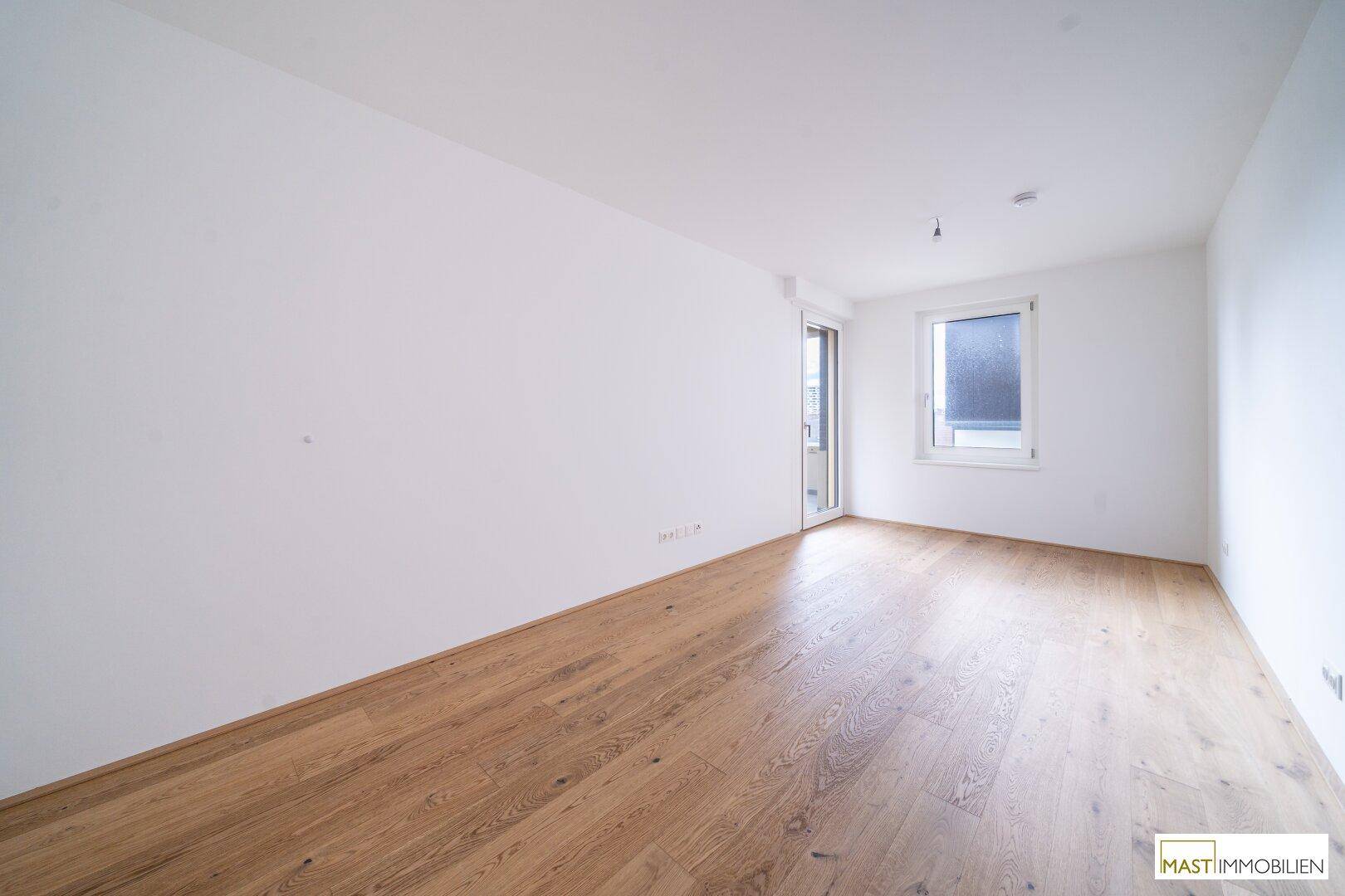 Freundlicher, leerer Raum mit hellem Parkettboden und einem Fenster, das für angenehmes Tageslicht sorgt.