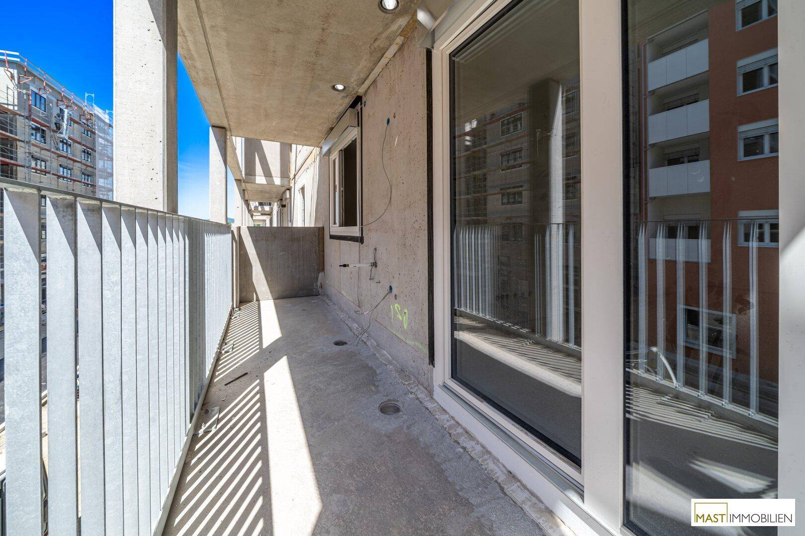 Langer, schmaler Balkon eines Neubaus mit Betonboden und Blick auf umliegende Gebäude und Baustellen.