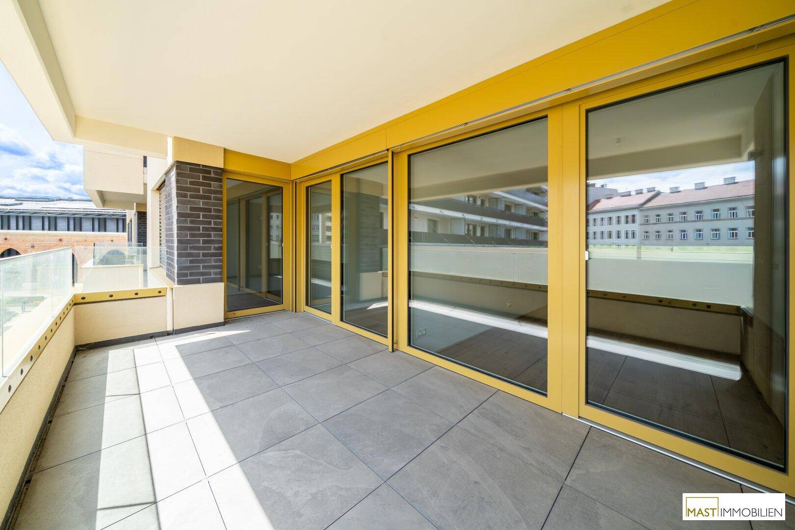 Geräumiger Balkon mit Fliesenboden, Glasgeländer und urbanem Ausblick.