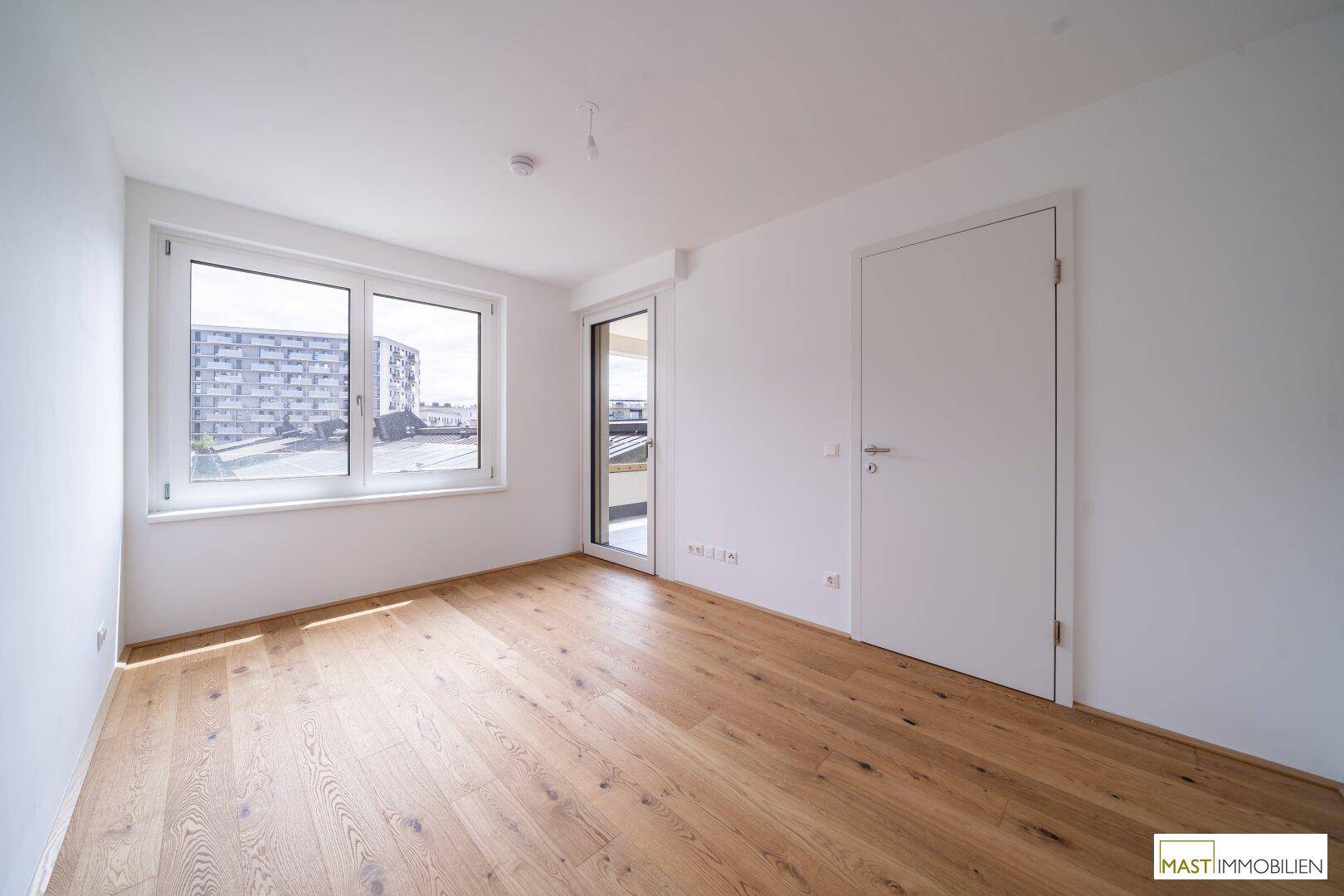 Leeres Zimmer mit Holzboden und großem Fenster, das viel Tageslicht hereinlässt.