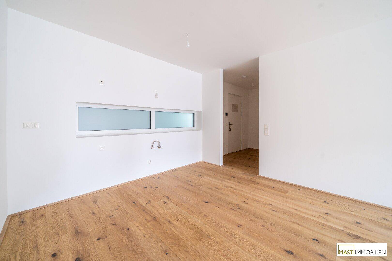 Leerer Küchenbereich mit Parkettboden, weißen Wänden und vorbereiteten Anschlüssen für eine Einbauküche.