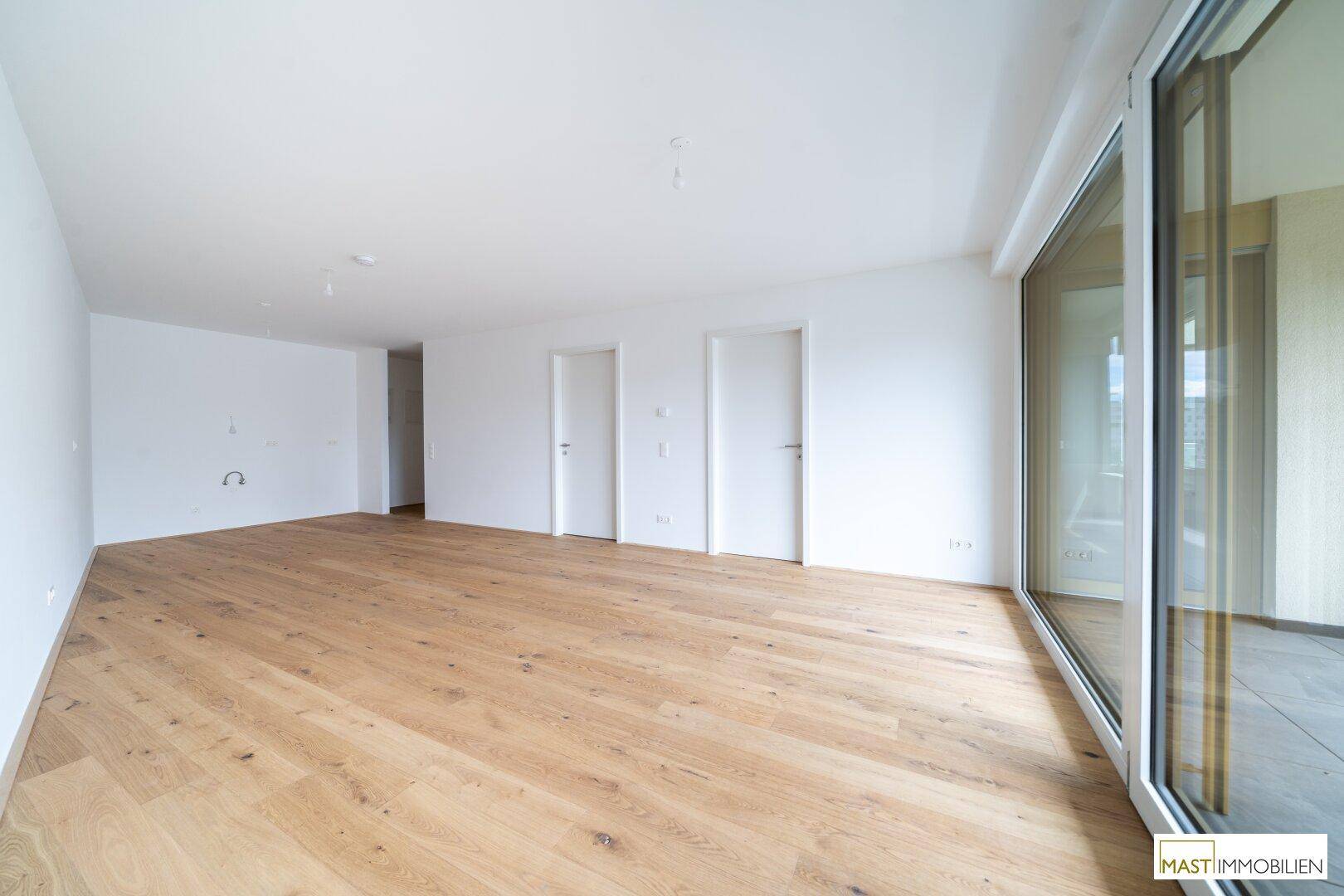Geräumiges, leeres Zimmer mit Holzboden und breiter Glasfront zum Außenbereich.