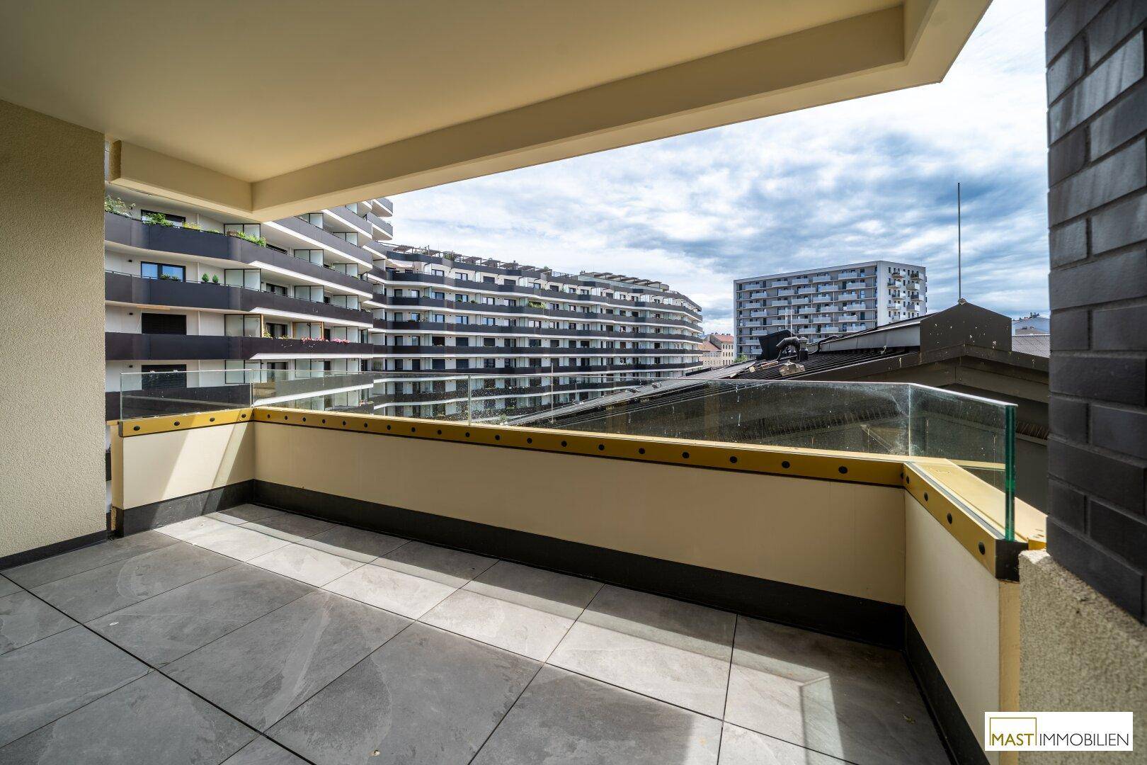Großer Balkon mit Glasgeländer und Fliesenboden, bietet weite Aussicht auf die Stadt.