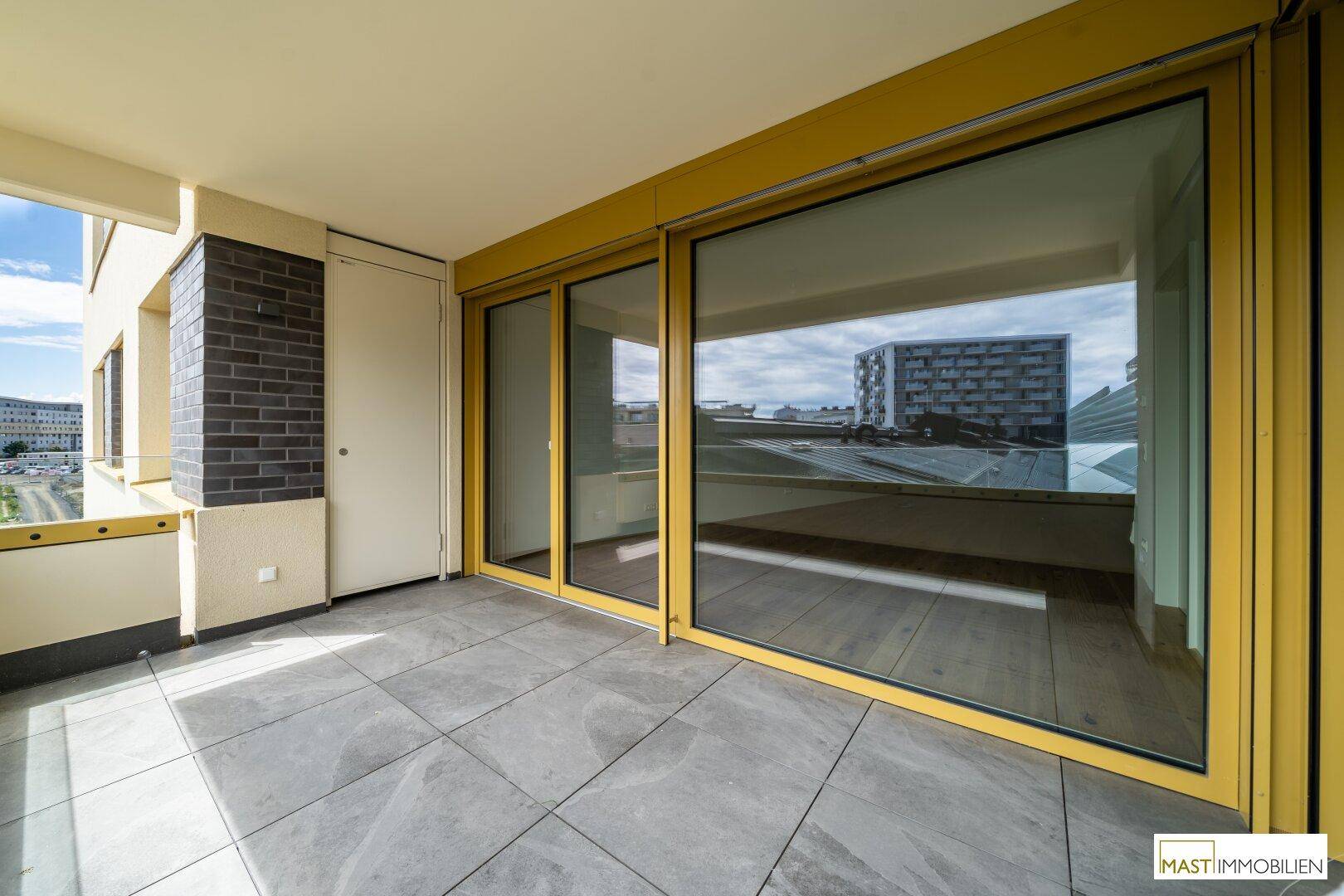 Geräumiger Balkon mit Fliesenboden und gelben Fensterrahmen, bietet Blick auf umliegende Gebäude.