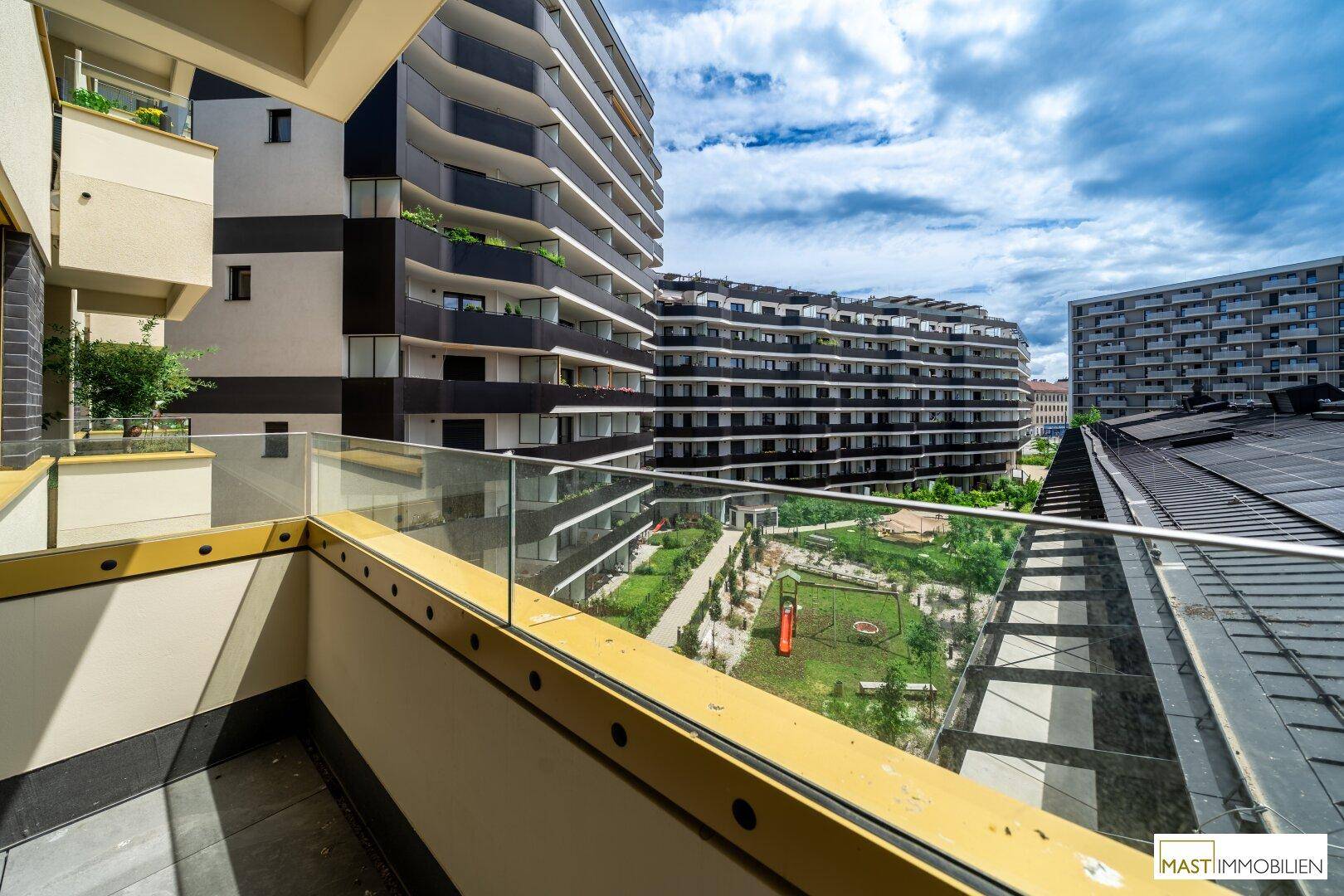 Balkon mit Glasgeländer und Blick auf moderne Gebäude sowie einen grünen Spielplatzbereich.