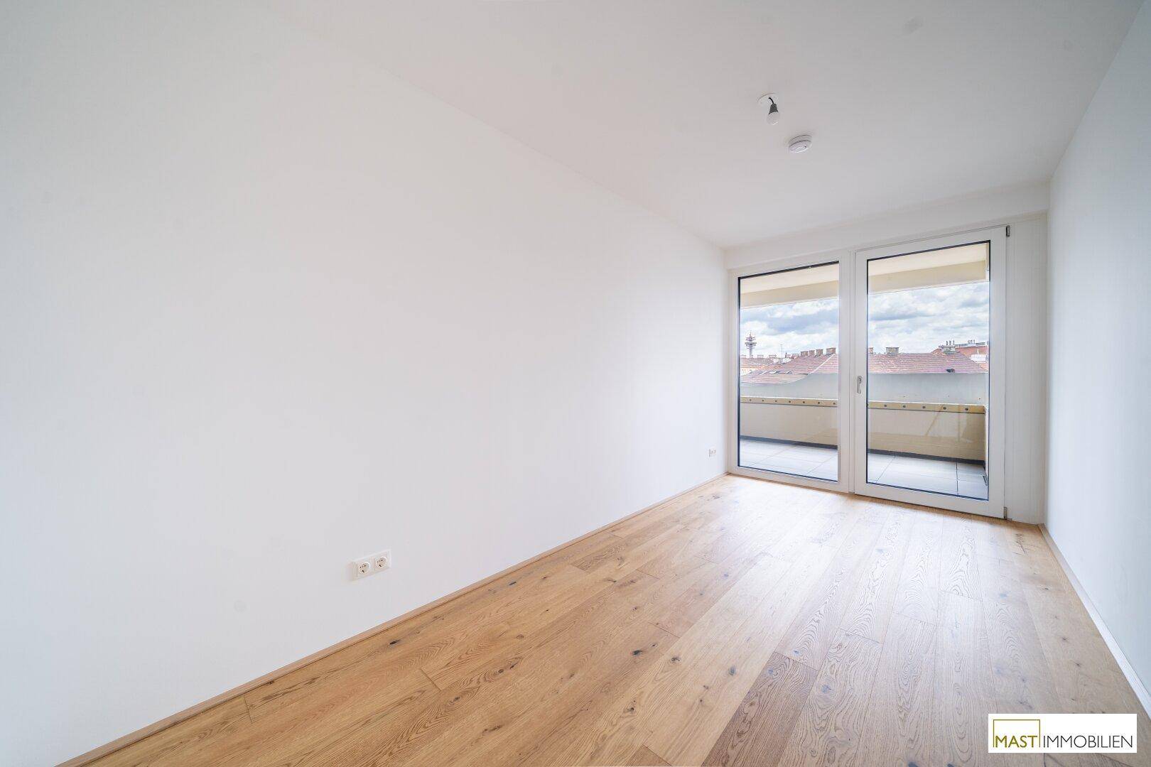 Geräumiges Zimmer mit hellem Holzparkett und großen Schiebetüren, die Zugang zu einem Balkon bieten.