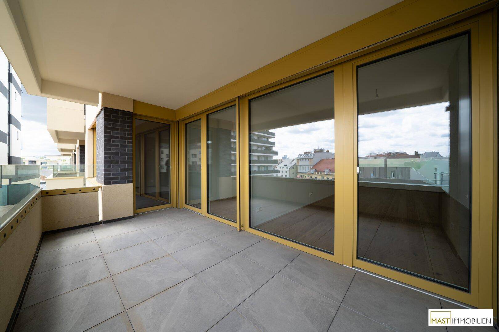 Geräumiger Balkon mit Fliesenboden und gelben Fensterrahmen, bietet einen weiten Blick über die Stadt.