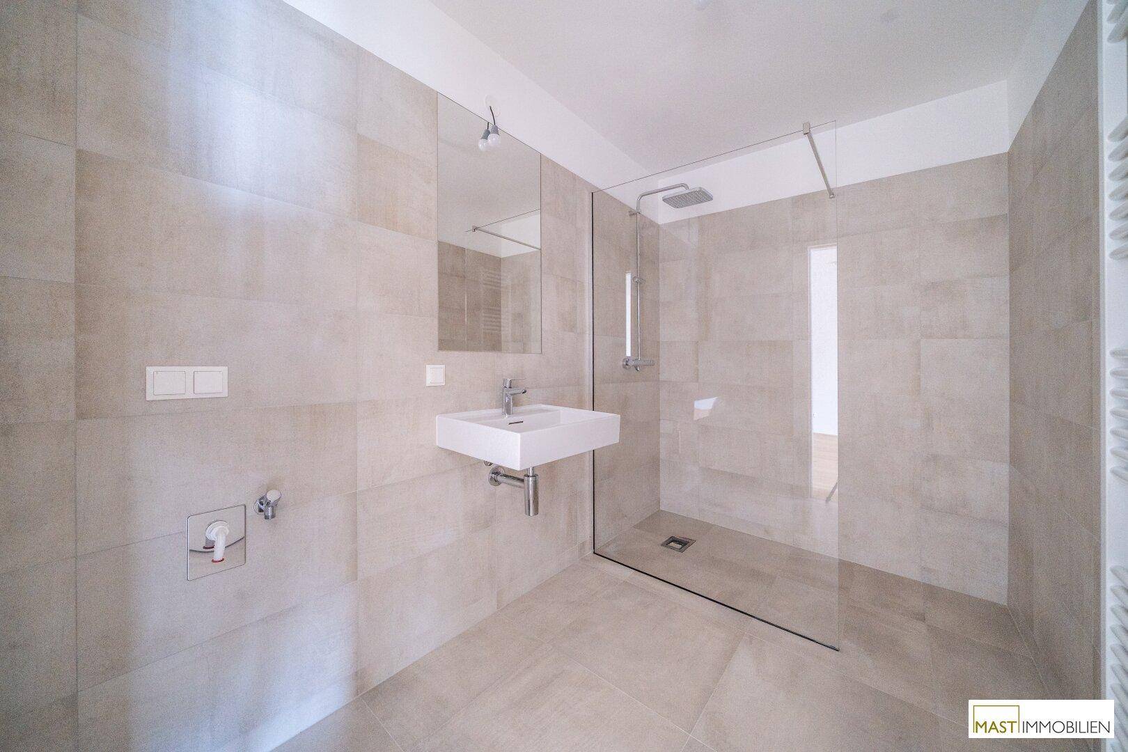 Modernes Badezimmer mit großen beigefarbenen Fliesen, ebenerdiger Dusche und Wandwaschbecken.