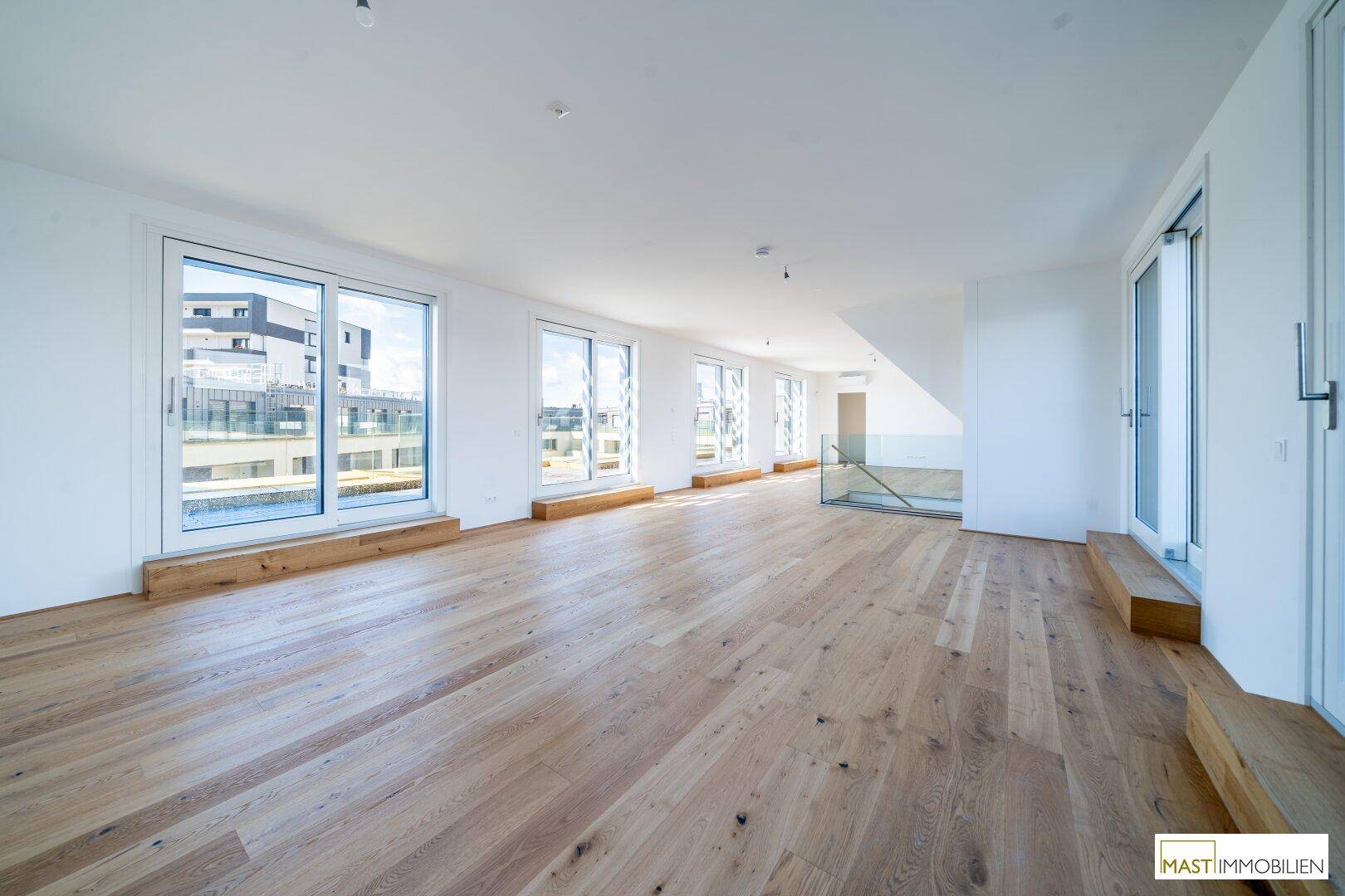 Geräumiges, helles Wohnzimmer mit Holzboden und großen Fenstern, die viel Tageslicht hereinlassen.