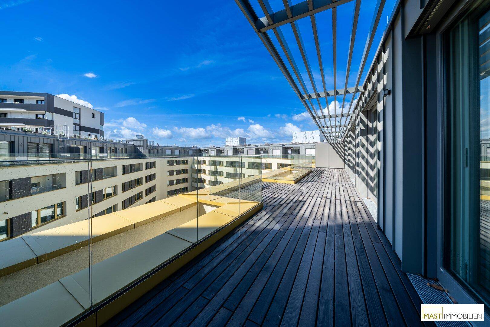 Weitläufige Dachterrasse mit Holzdielen und Glasgeländer, umgeben von modernen Gebäuden.