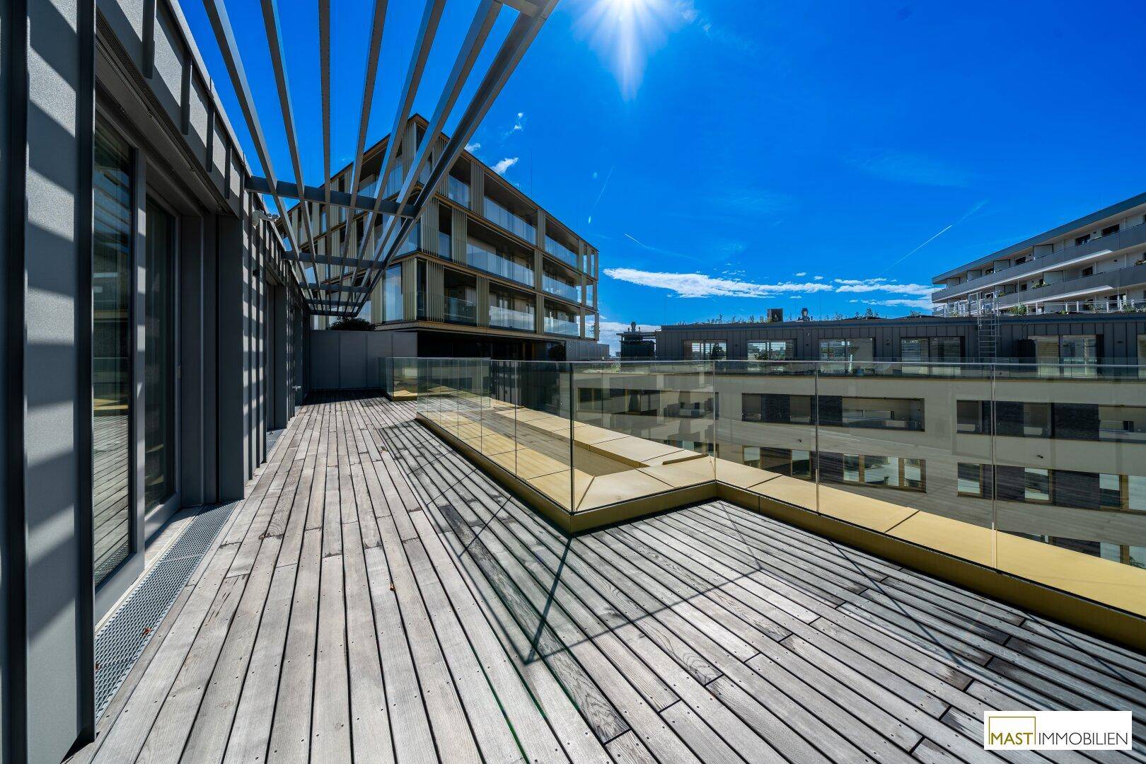 Große Dachterrasse mit Holzboden, Glasgeländer und Blick auf moderne Gebäude.
