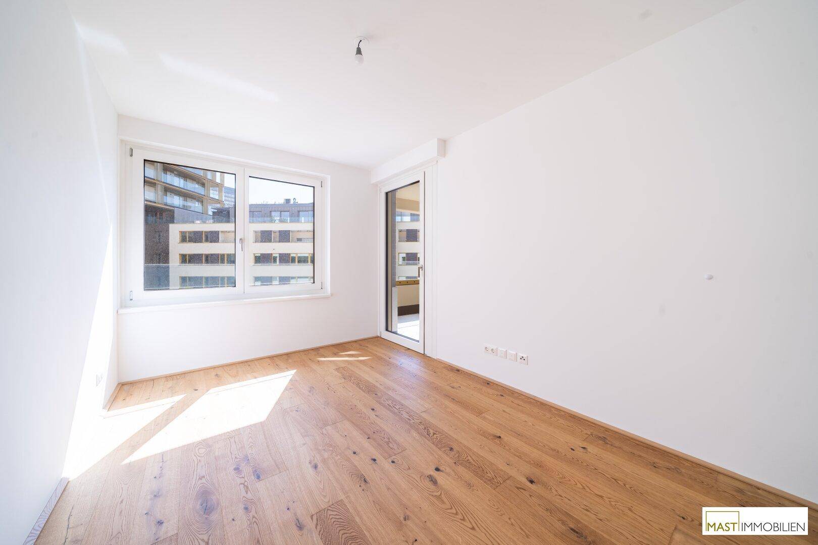 Sonniger Raum mit Parkettboden und großem Fenster, das viel Tageslicht hereinlässt.