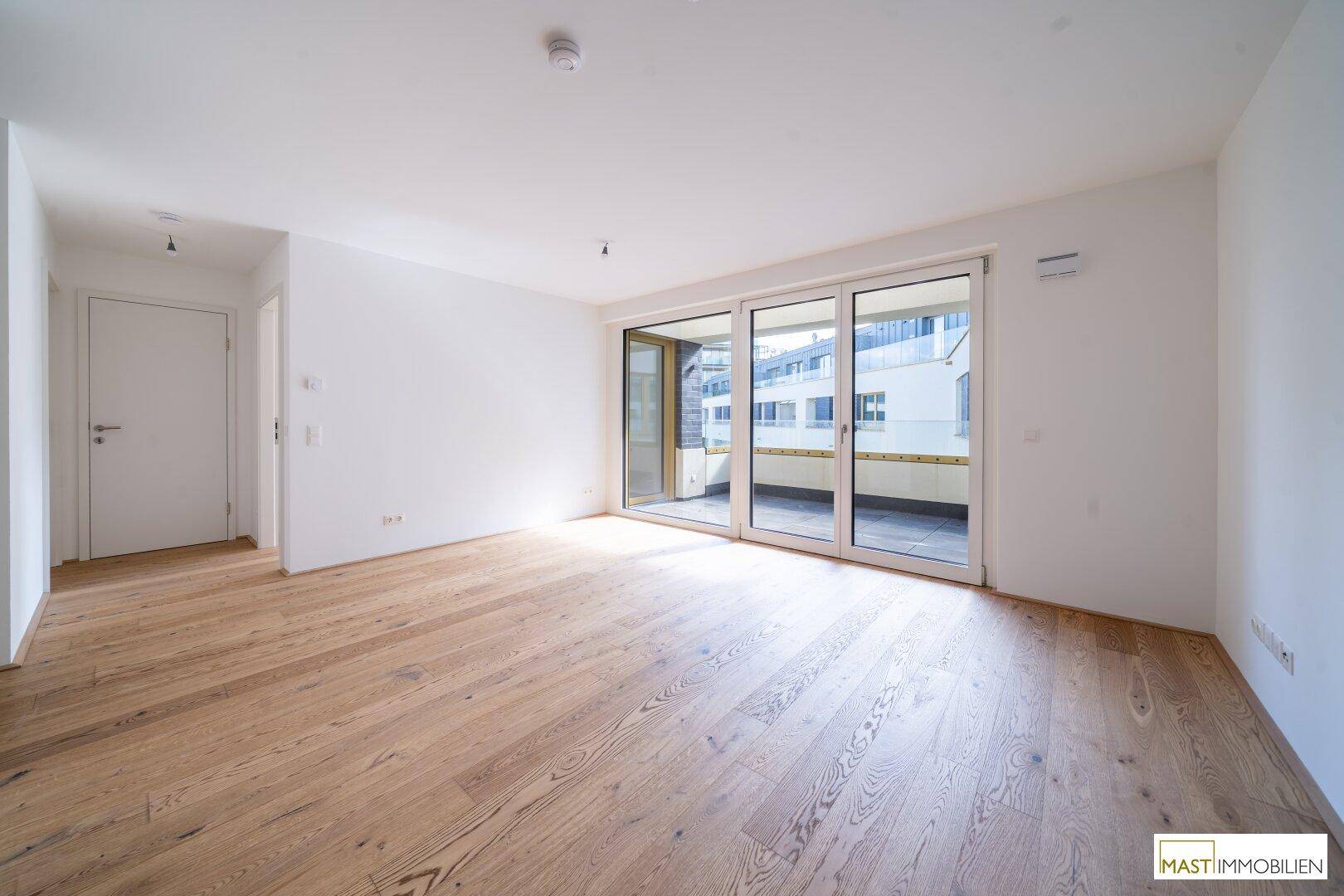 Geräumiges Wohnzimmer mit Holzboden und Zugang zu einem Balkon, bietet viel Tageslicht.