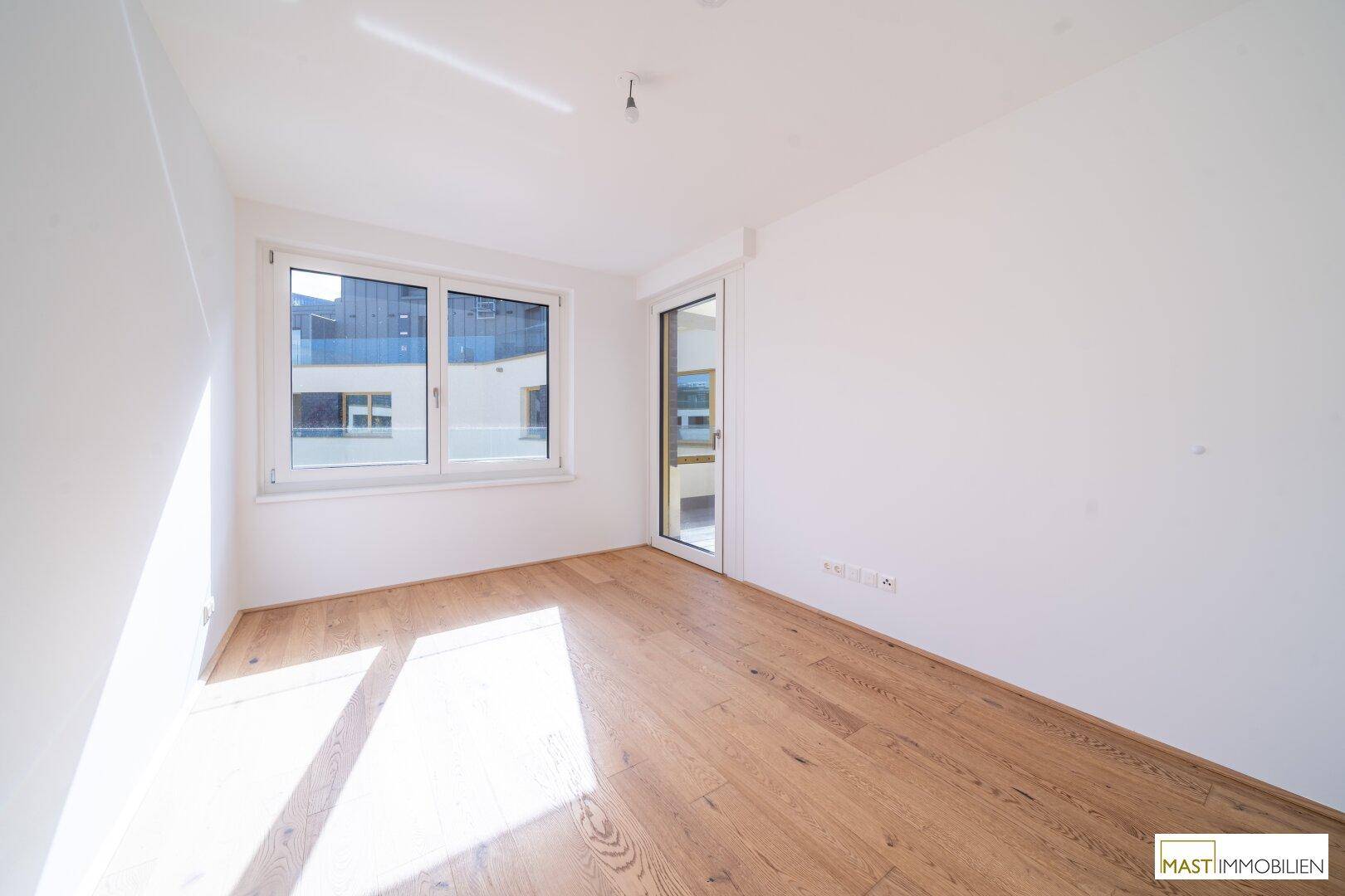 Sonniger Raum mit Holzboden und großem Fenster, ideal als Schlafzimmer oder Arbeitszimmer.